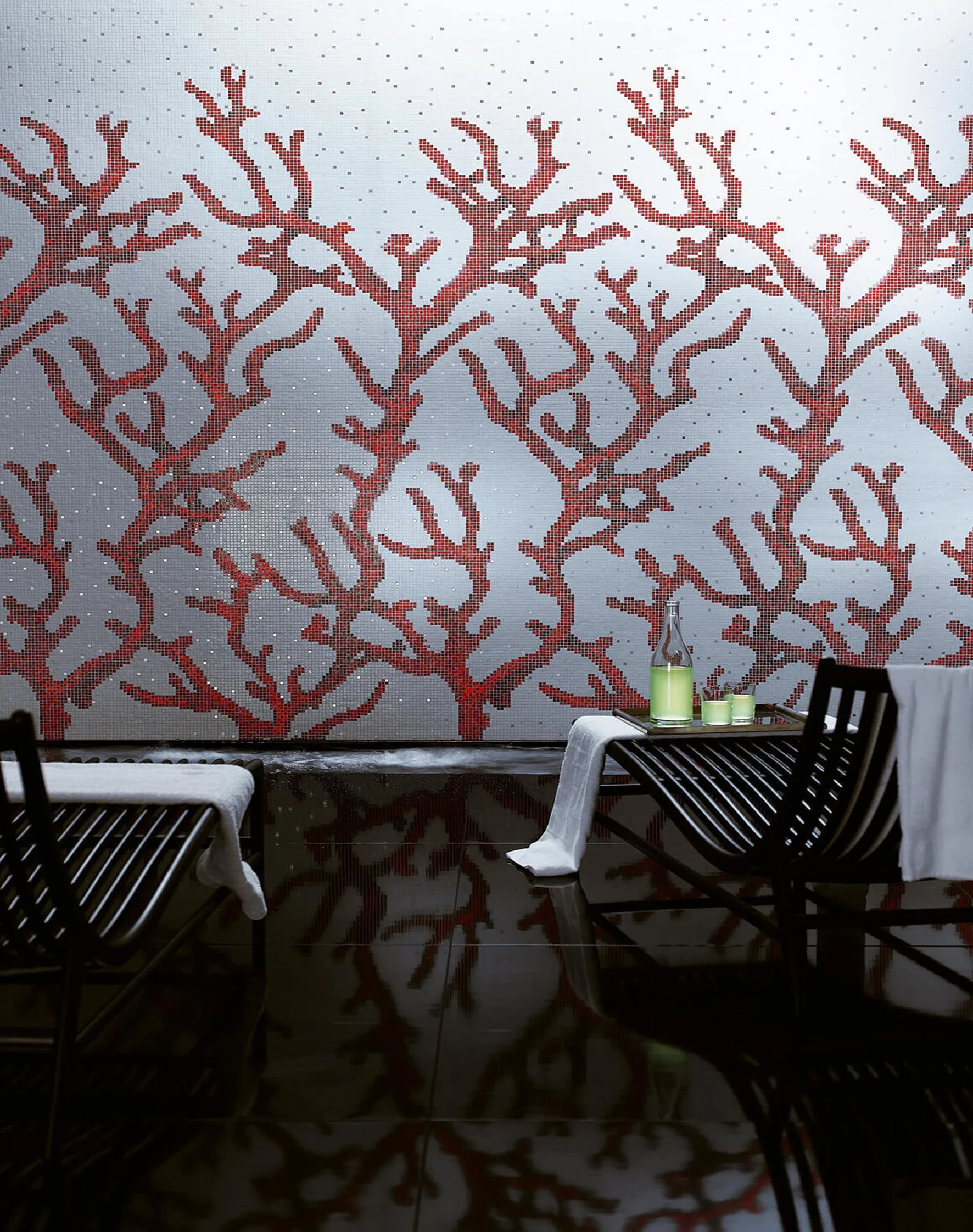 Sillas de madera negras con mantas blancas, una bandeja con una botella y vasos de color verde, un fondo con pared decorativa de coral rojo y brillo plateado.