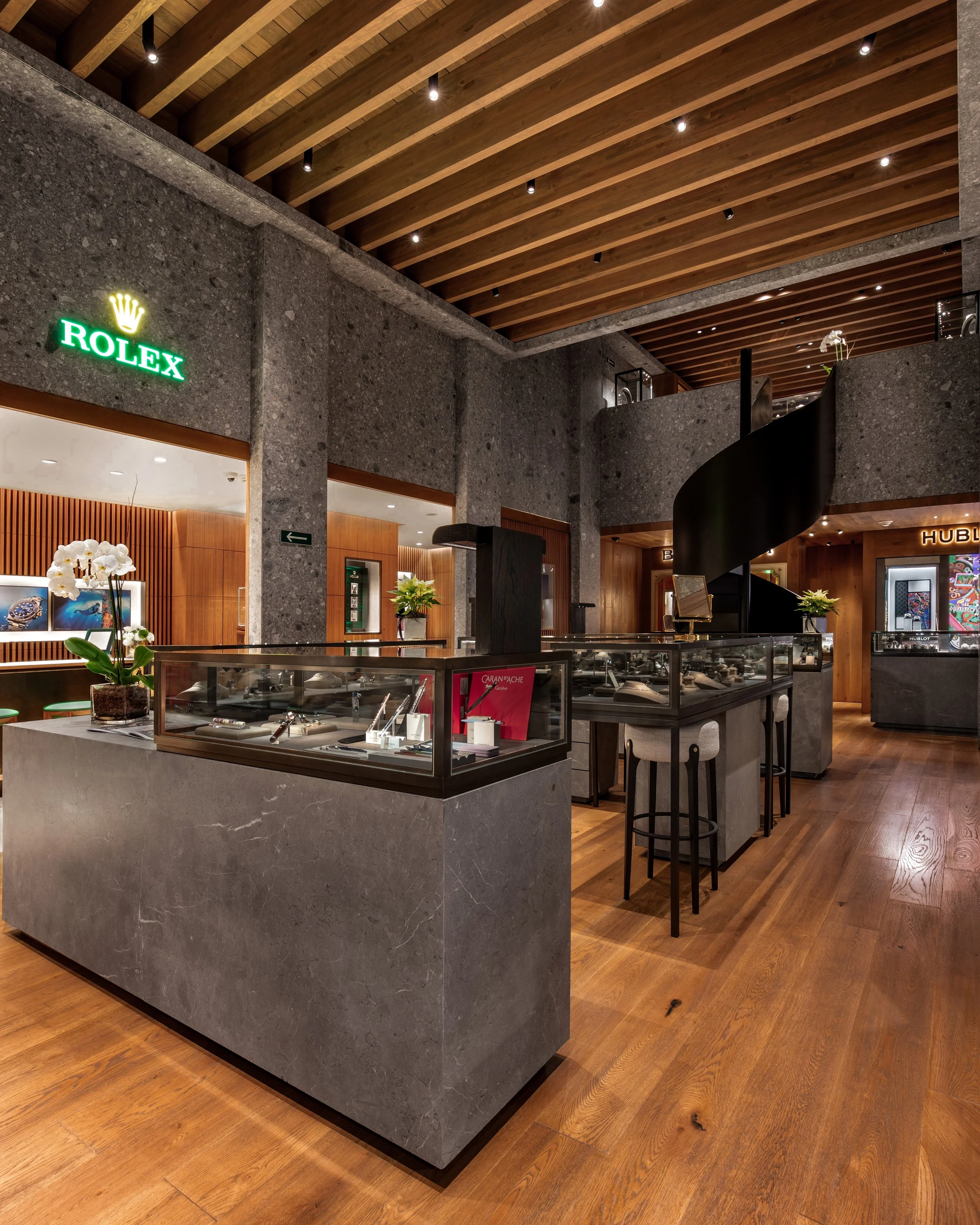 Vista interior de una tienda de lujo con exhibidores de relojes en vidrio, plantas ornamentales, paredes de piedra y piso de madera, con cartel de Rolex iluminado en verde.