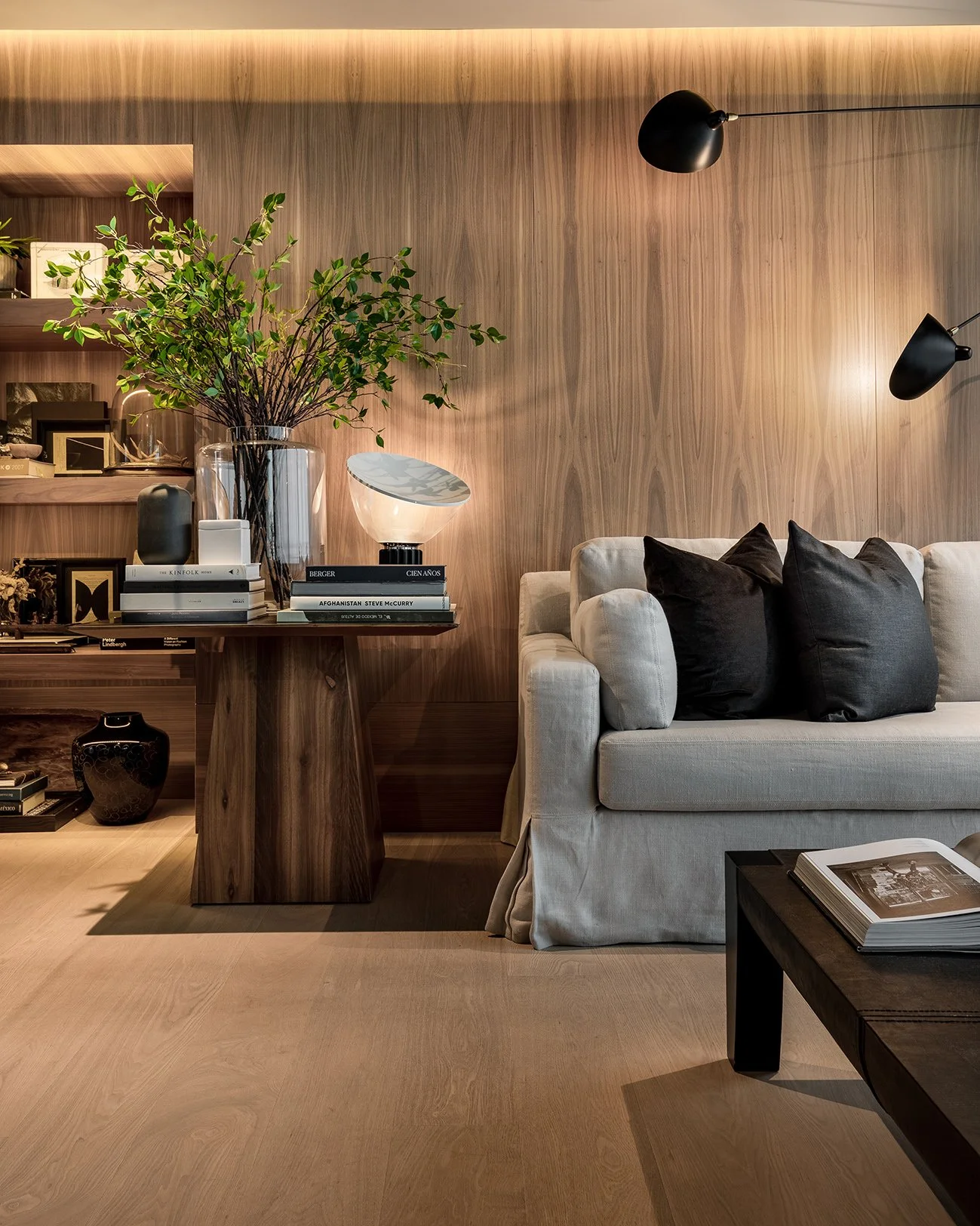 Sala de estar con pared de madera, sofá beige con cojines oscuros, mesa de madera con libro abierto, y estantería con libros y decoraciones, lámparas negras y jarrón con plantas verdes.