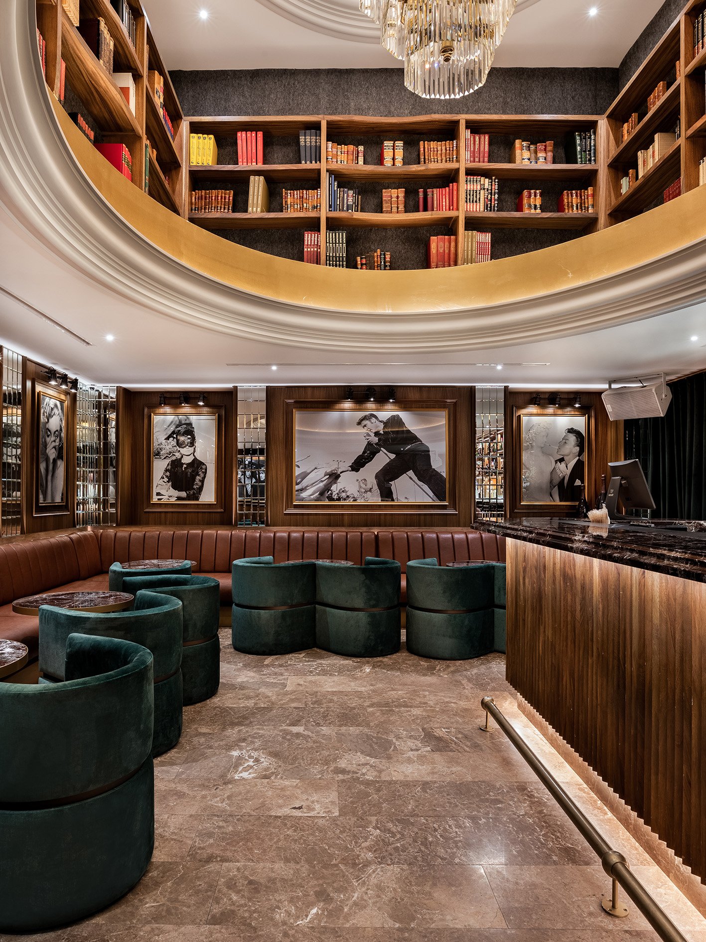 Lobby elegante con estanterías llenas de libros en la segunda planta, paredes decoradas con fotografías en blanco y negro, área de sillones verdes y un bar de madera con mármol negro.