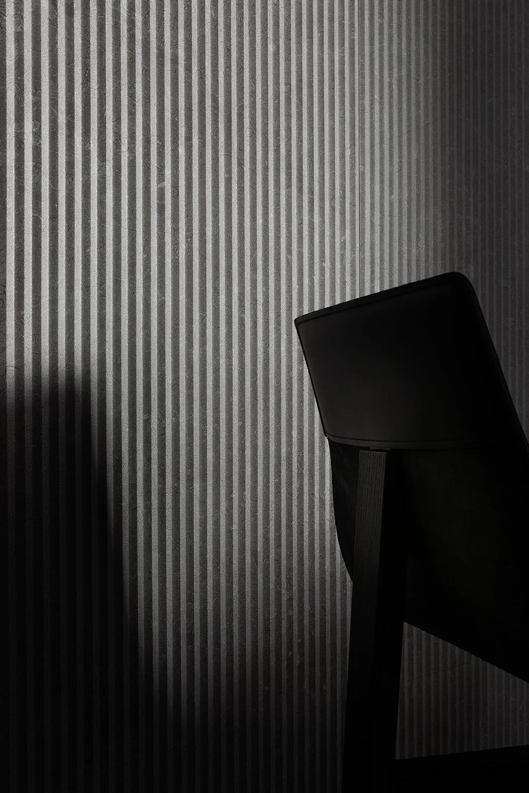 Una silla negra junto a una pared de fondo gris con líneas verticales.