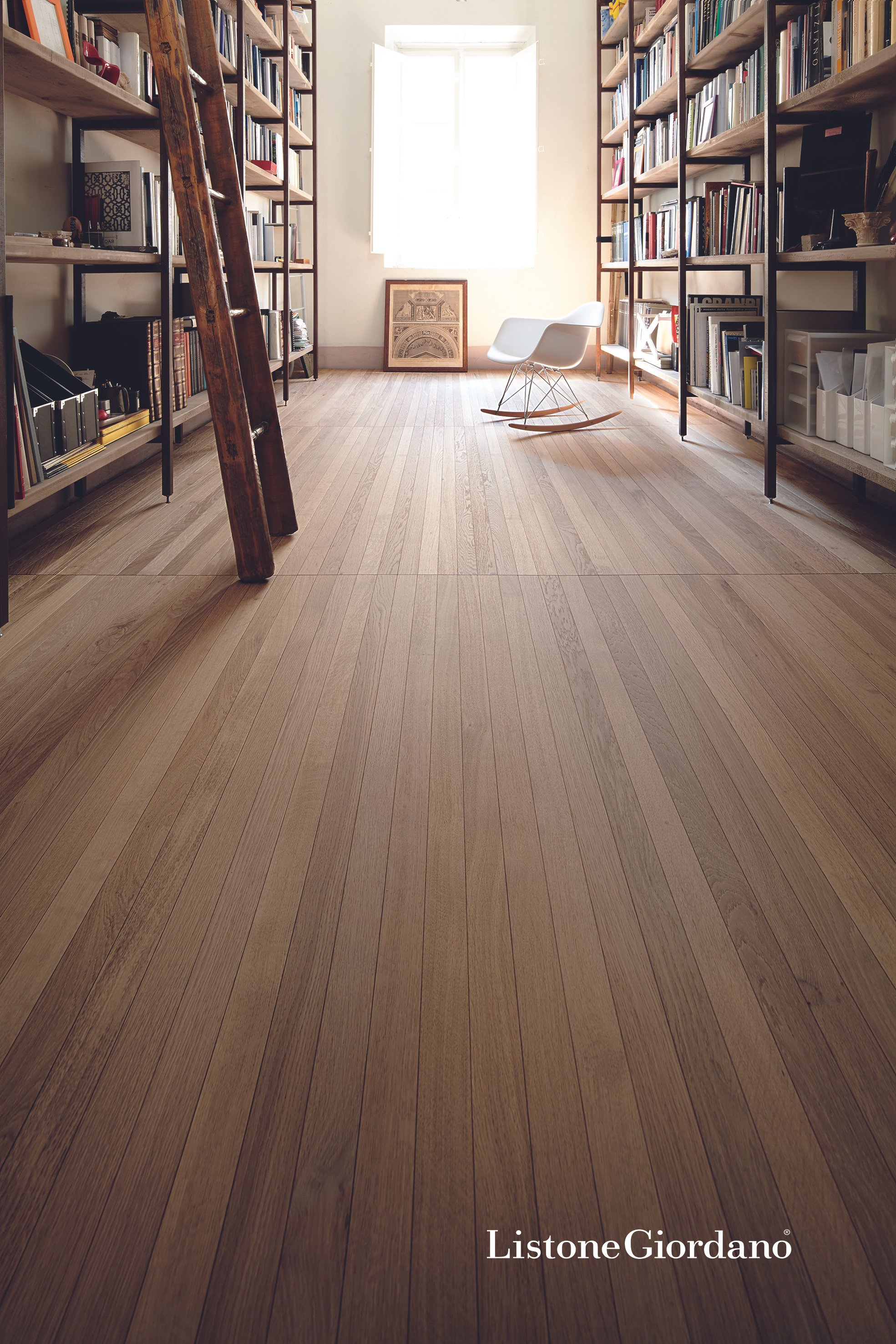 Sala de lectura o estudio con estanterías llenas de libros, una silla blanca de mecedora, una ventana grande y cuadros de arte en el fondo. Piso de madera clara y ambiente luminoso.