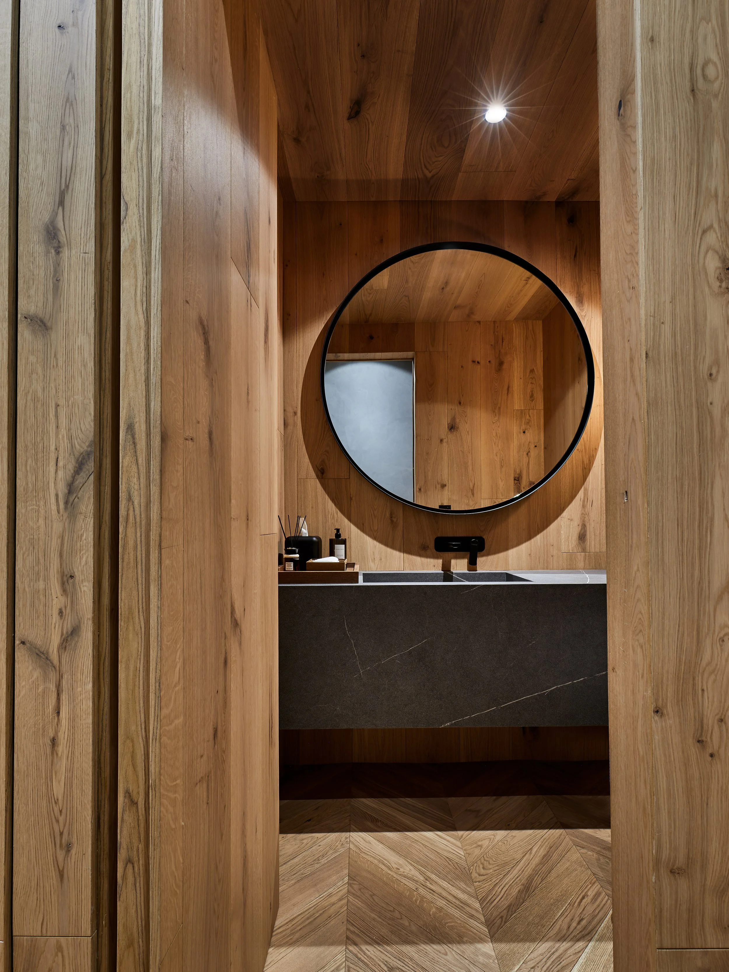 Espejo circular sobre un lavabo de piedra en un baño con paredes y suelo de madera.