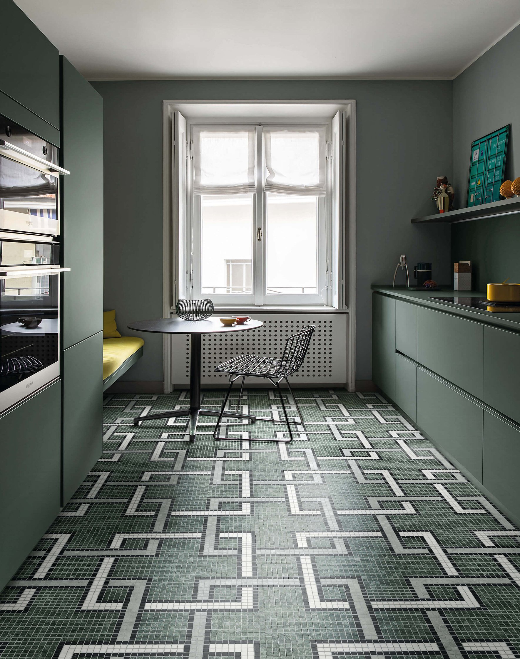 Cocina moderna con gabinetes verdes y piso de mosaico, ventana grande con cortinas blancas, mesa redonda y silla de alambre