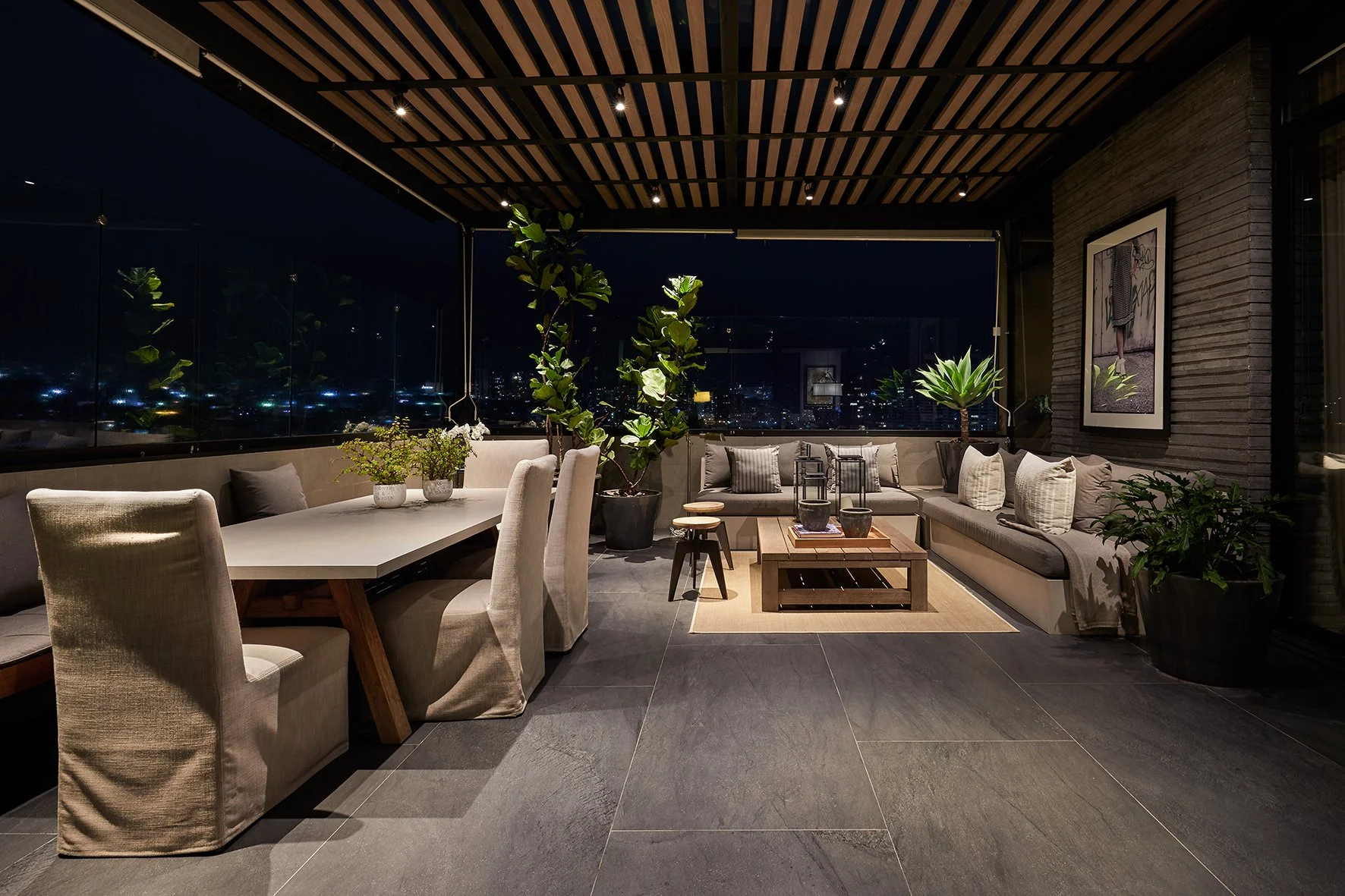 Terraza moderna con iluminación nocturna, sofá de esquina con cojines, mesa de centro de madera, comedor con sillas tapizadas, plantas grandes y cuadros decorativos en las paredes.