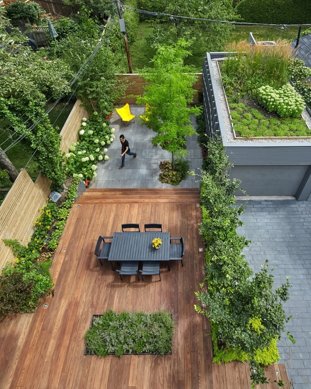 Vista aérea de un patio con área de comedor con mesa y sillas negras y plantas verdes, dividido en una zona de madera y otra de piedra con árboles y arbustos