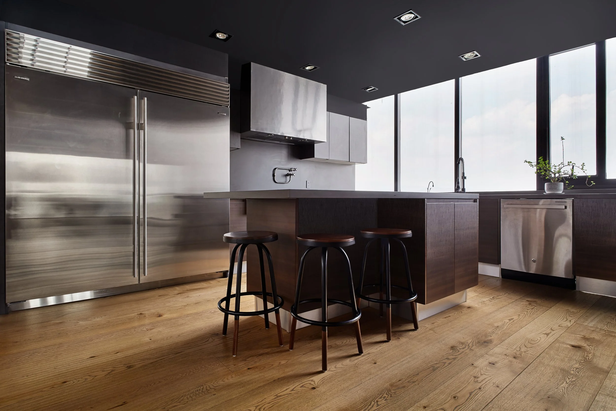 Cocina moderna con nevera de acero inoxidable, isla con taburetes, encimera blanca, ventanas grandes y plantas decorativas.