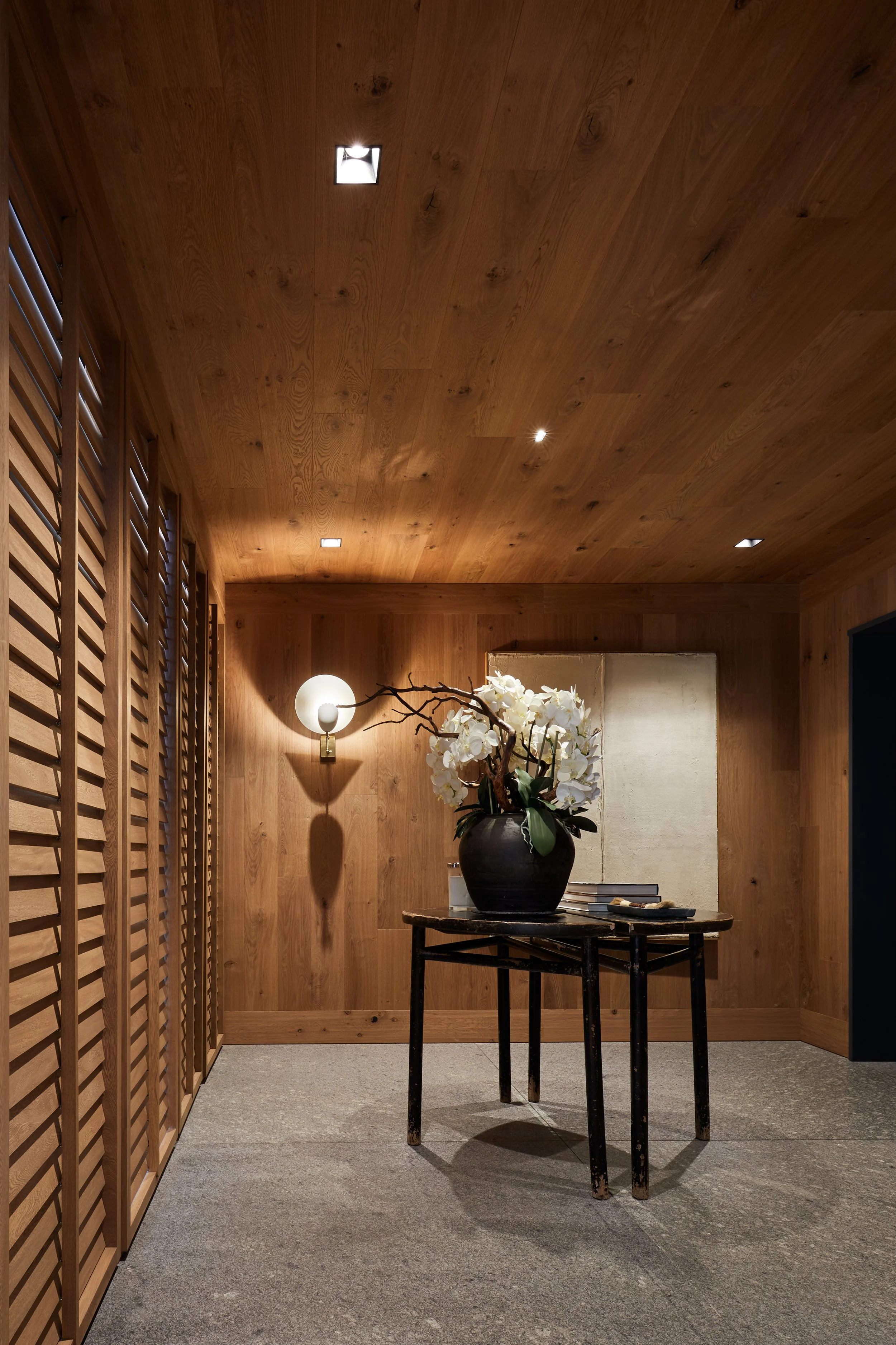 Sala con paredes y techo de madera, lámpara de pared, mesa negra con jarrón de flores blancas y libros en una habitación elegante y moderna.