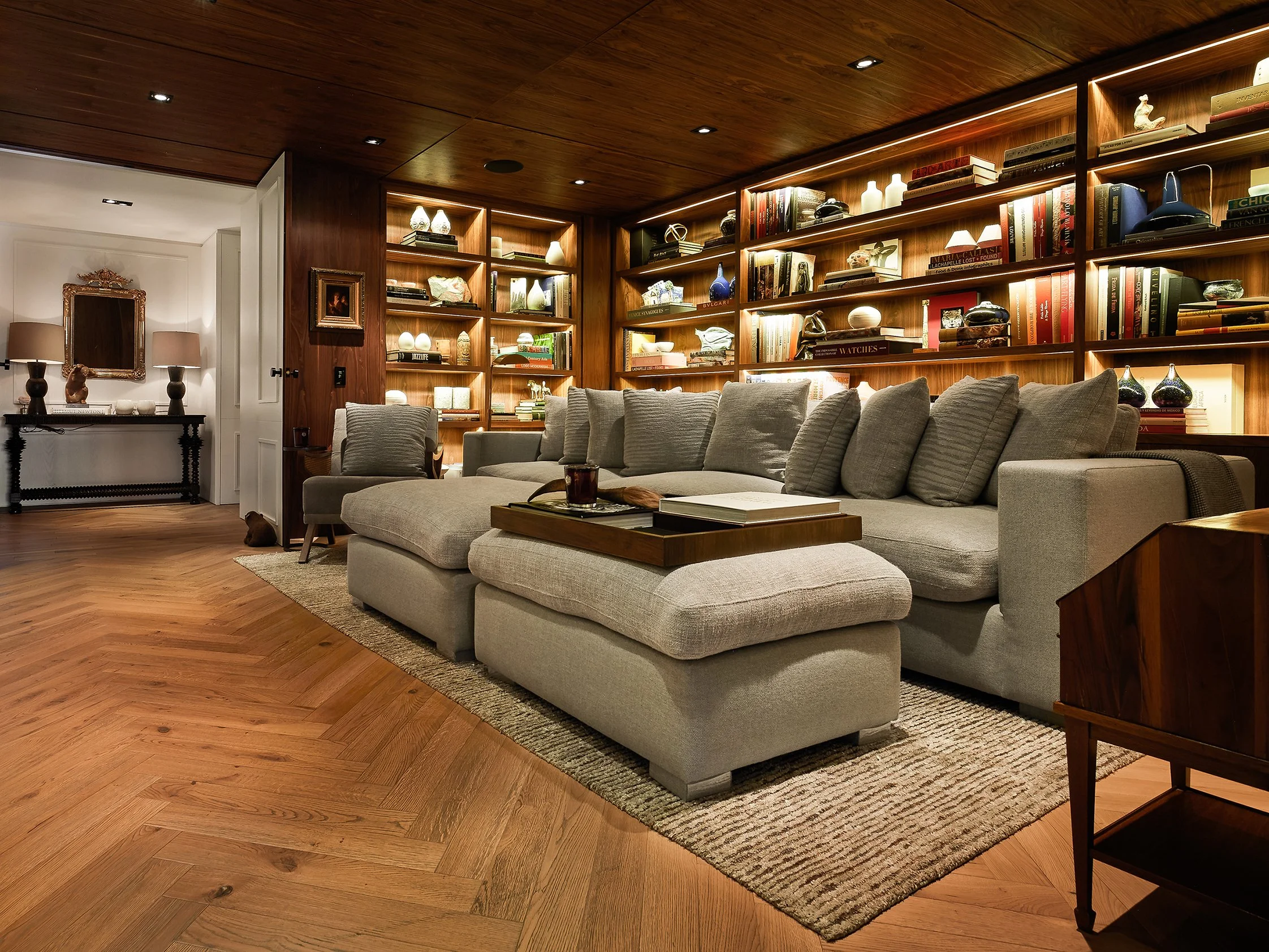 Sala de estar con sofá gris, estanterías de madera llenas de libros y objetos decorativos, suelo de madera y paredes blancas. Hay lámparas y accesorios decorativos en las estanterías.