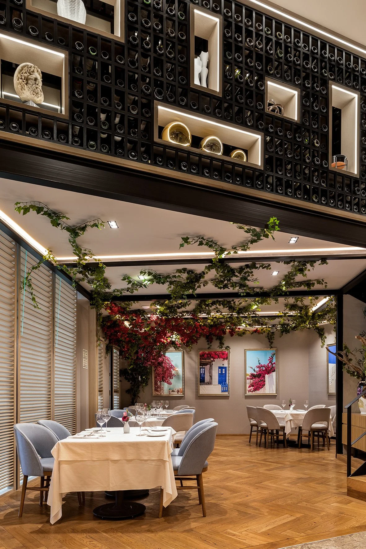 Interior de restaurante elegante con mesas con manteles blancos, copas y vajilla, decorado con plantas y cuadros en las paredes, con una estantería de vino en la parte superior.