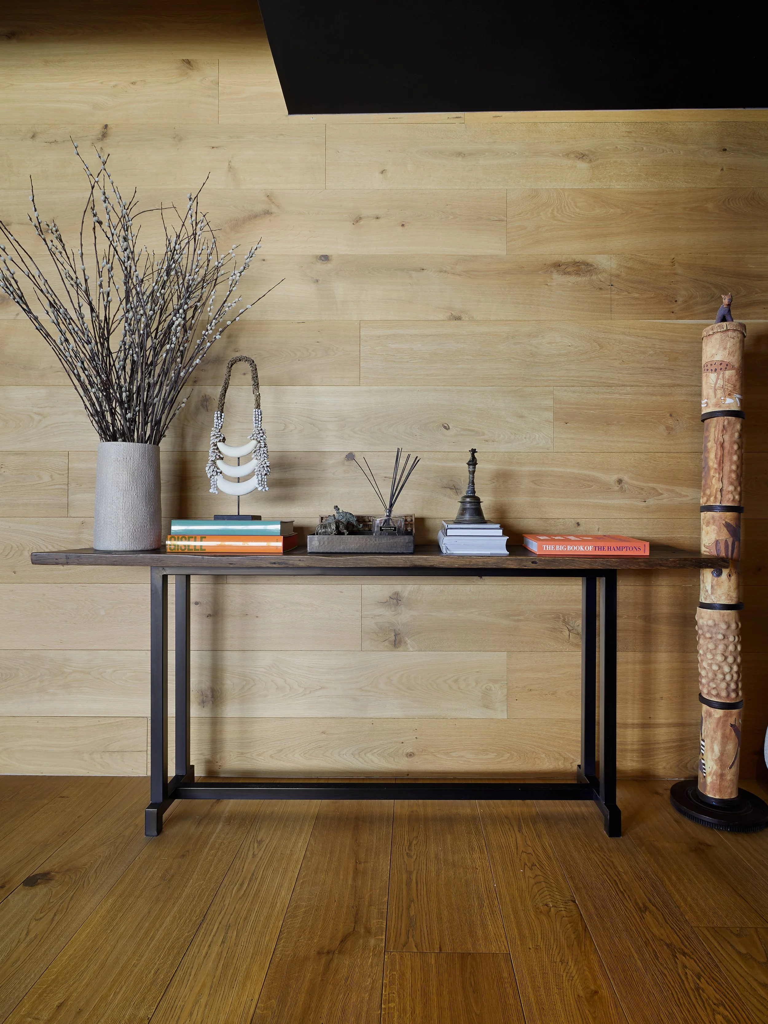 Una mesa de madera con marco de metal negro, con diversas decoraciones y libros, sobre una pared de madera clara. A la izquierda, un jarrón con ramas secas. A la derecha, un colgante decorativo y a un lado, un tubo decorativo de madera.