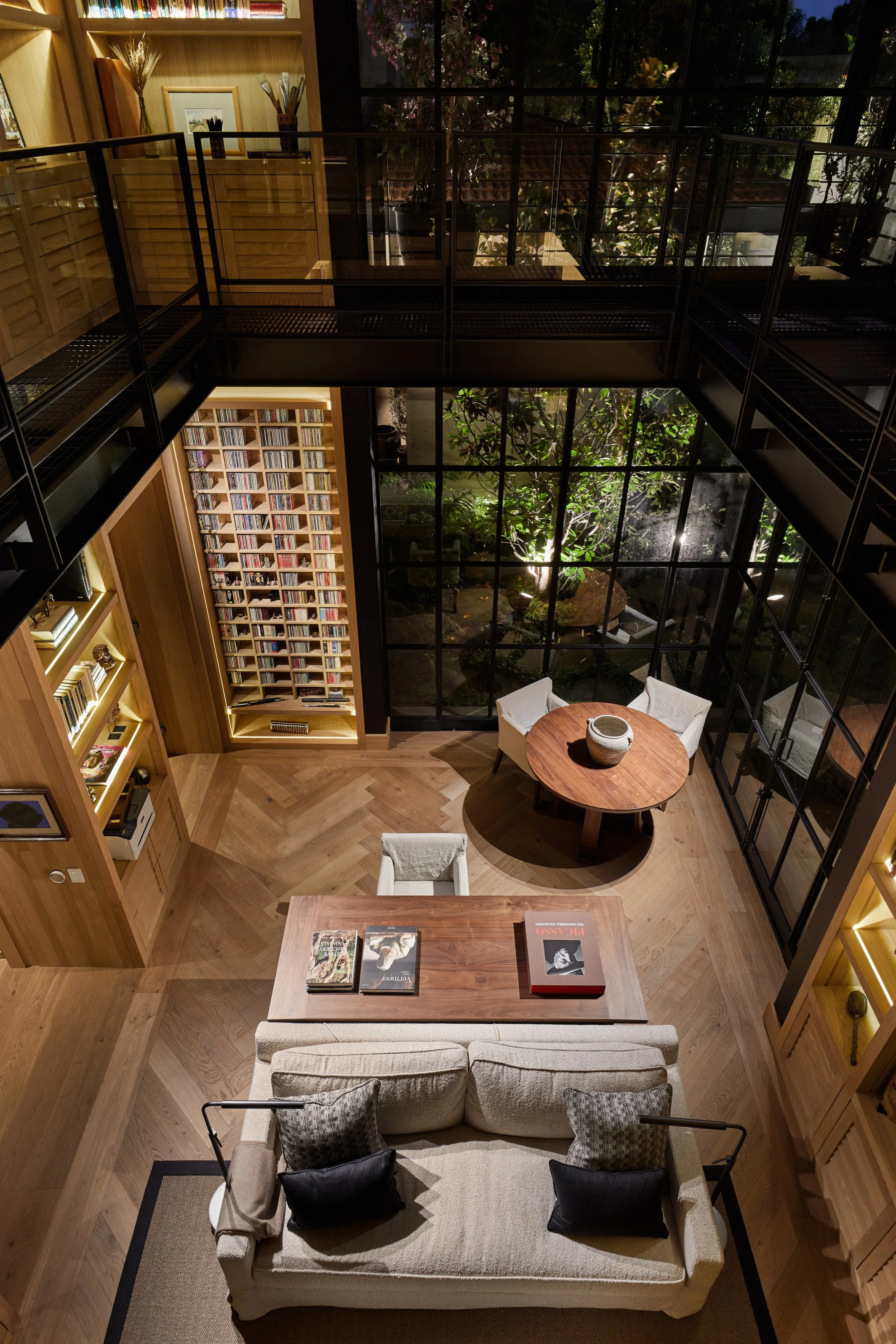 Vista de interior de una sala de lectura con paredes de ladrillo, estanterías llenas de libros, un sofá, una mesa de comedor, y ventanas grandes que muestran un jardín con plantas verdes.