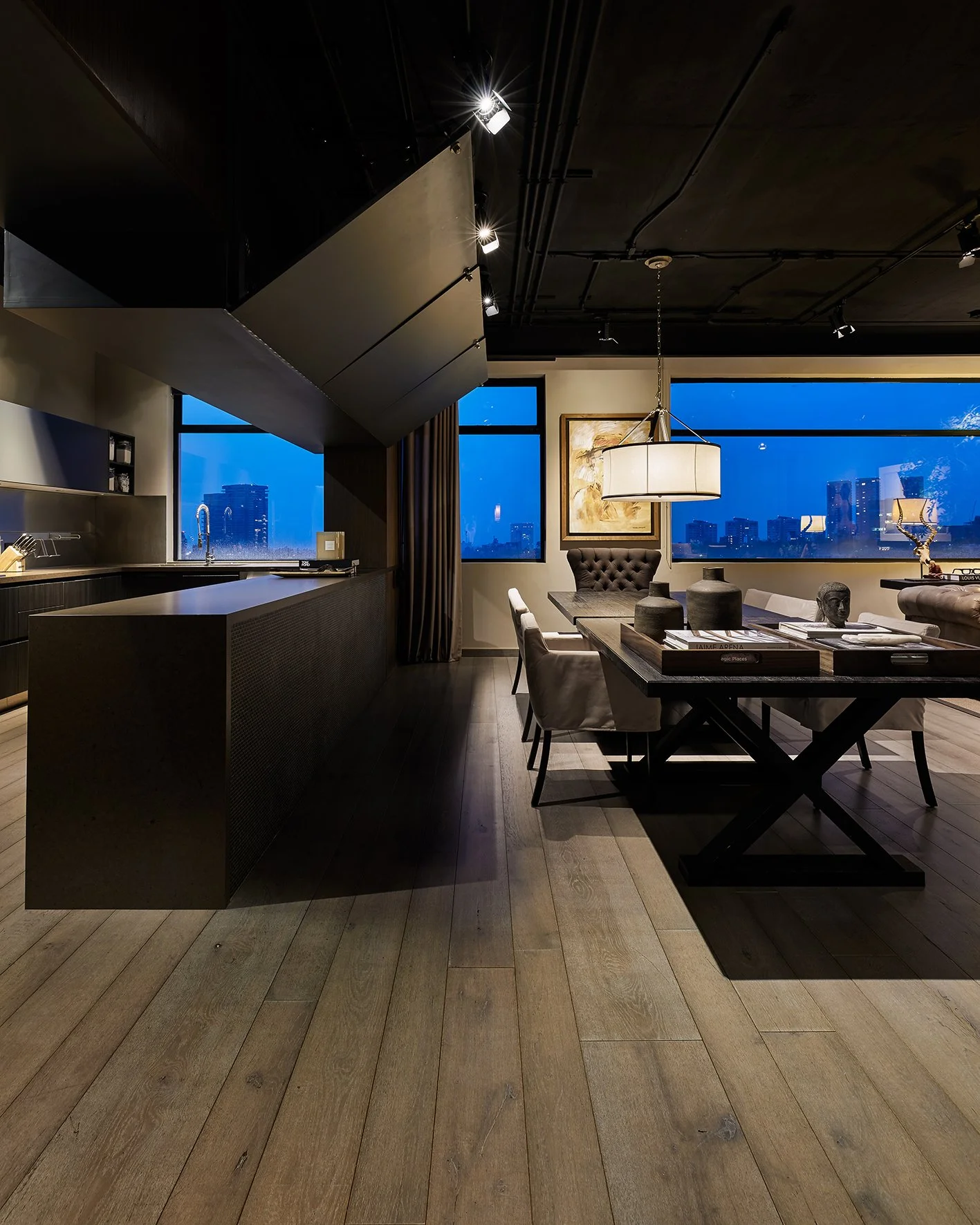 Interior de una sala moderna con ventanas grandes que muestran una ciudad al atardecer, con una mesa de comedor, sillas, luminaria y decoración en tonos neutros.