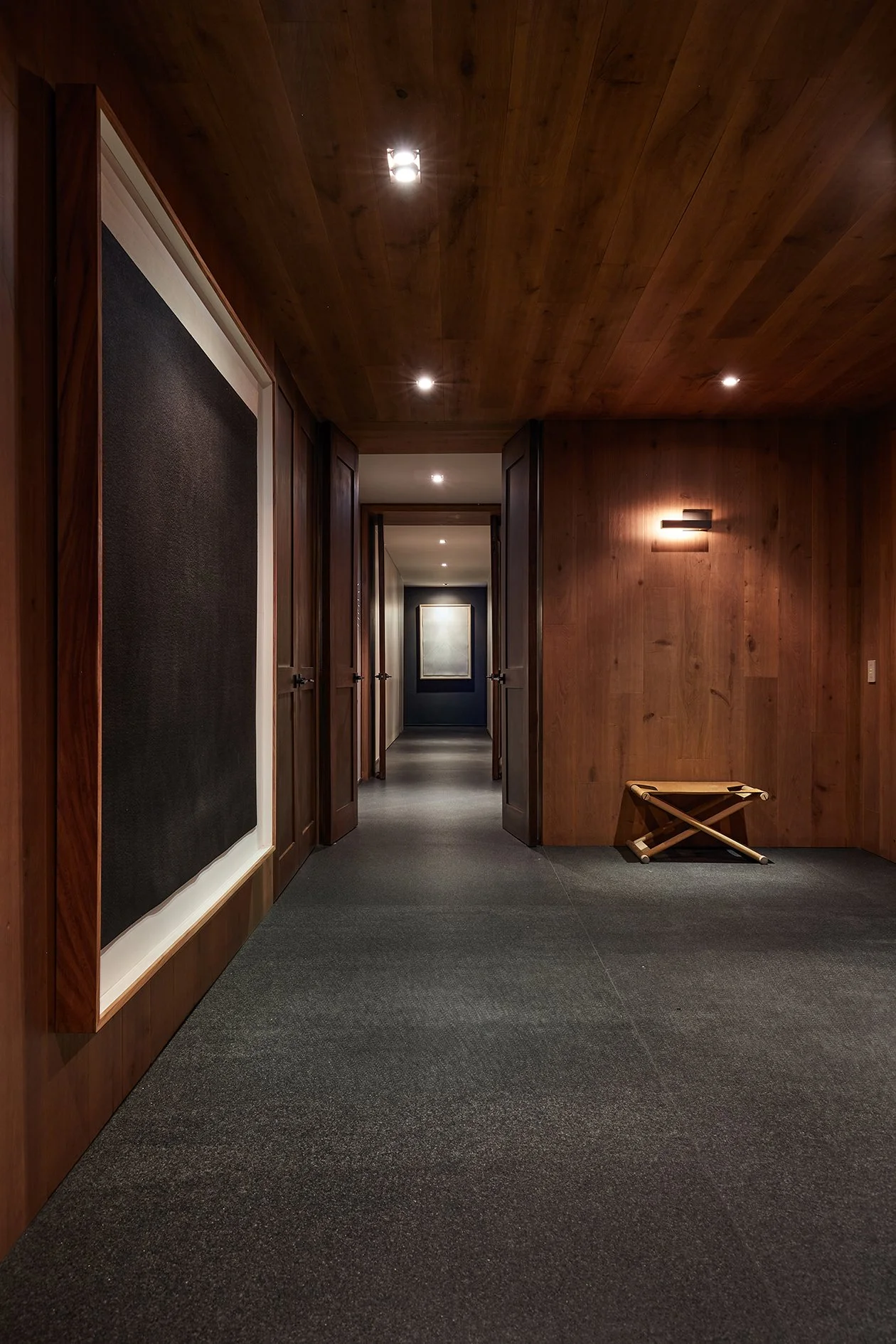 Pasillo con paredes y techo de madera, con varias puertas a los lados, una obra de arte en la pared y una silla plegable de madera en el lado derecho.