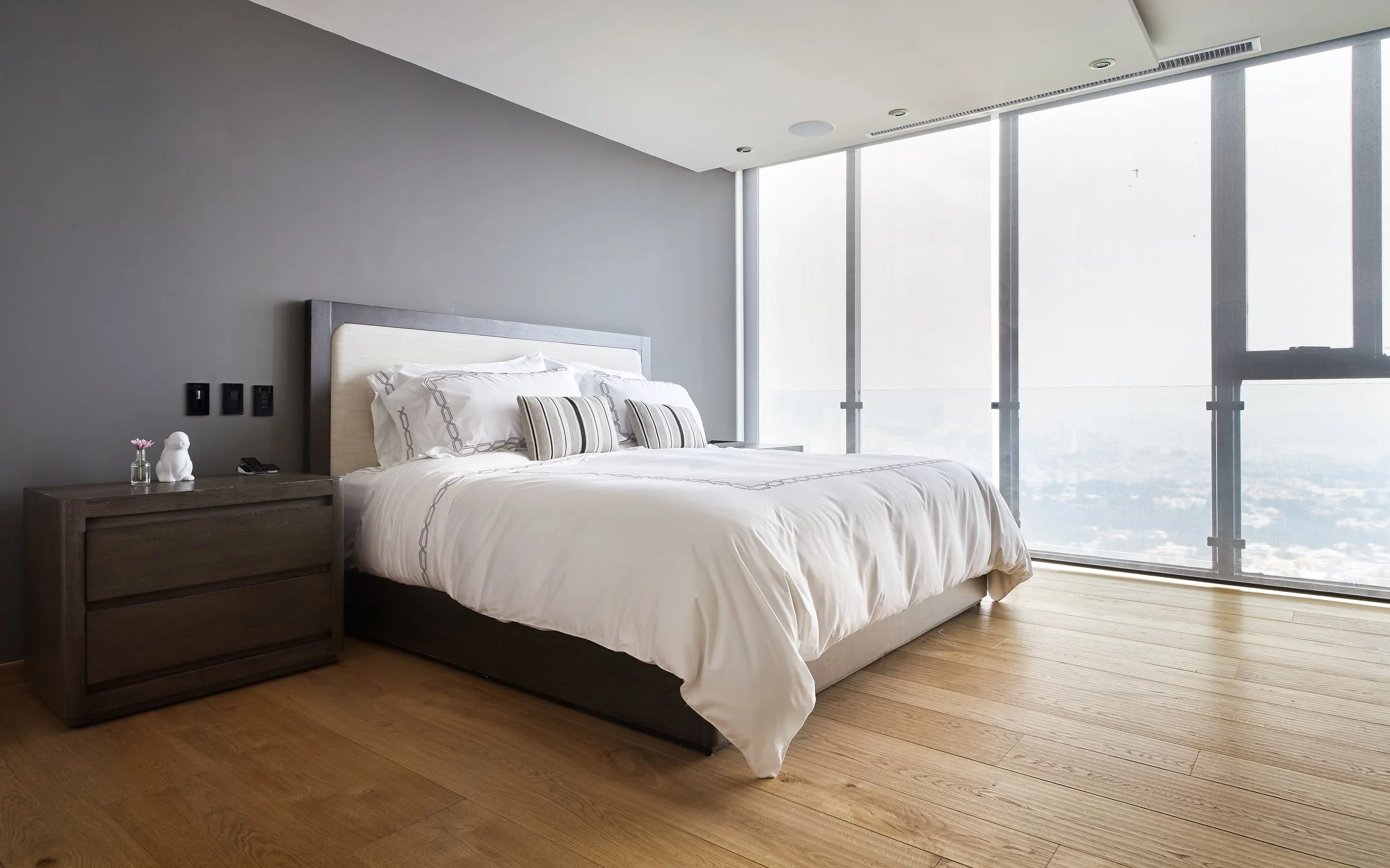 Habitación moderna con cama grande, ropa de cama blanca, almohadas a rayas y mesita de noche de madera oscura junto a una pared gris. Ventanas grandes con vista a la ciudad.