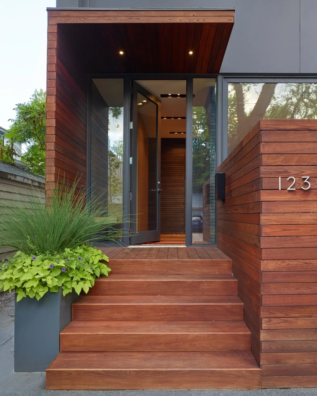 Entrada moderna de casa con escalones de madera, puerta de vidrio y paredes de madera, con plantas verdes a la izquierda.