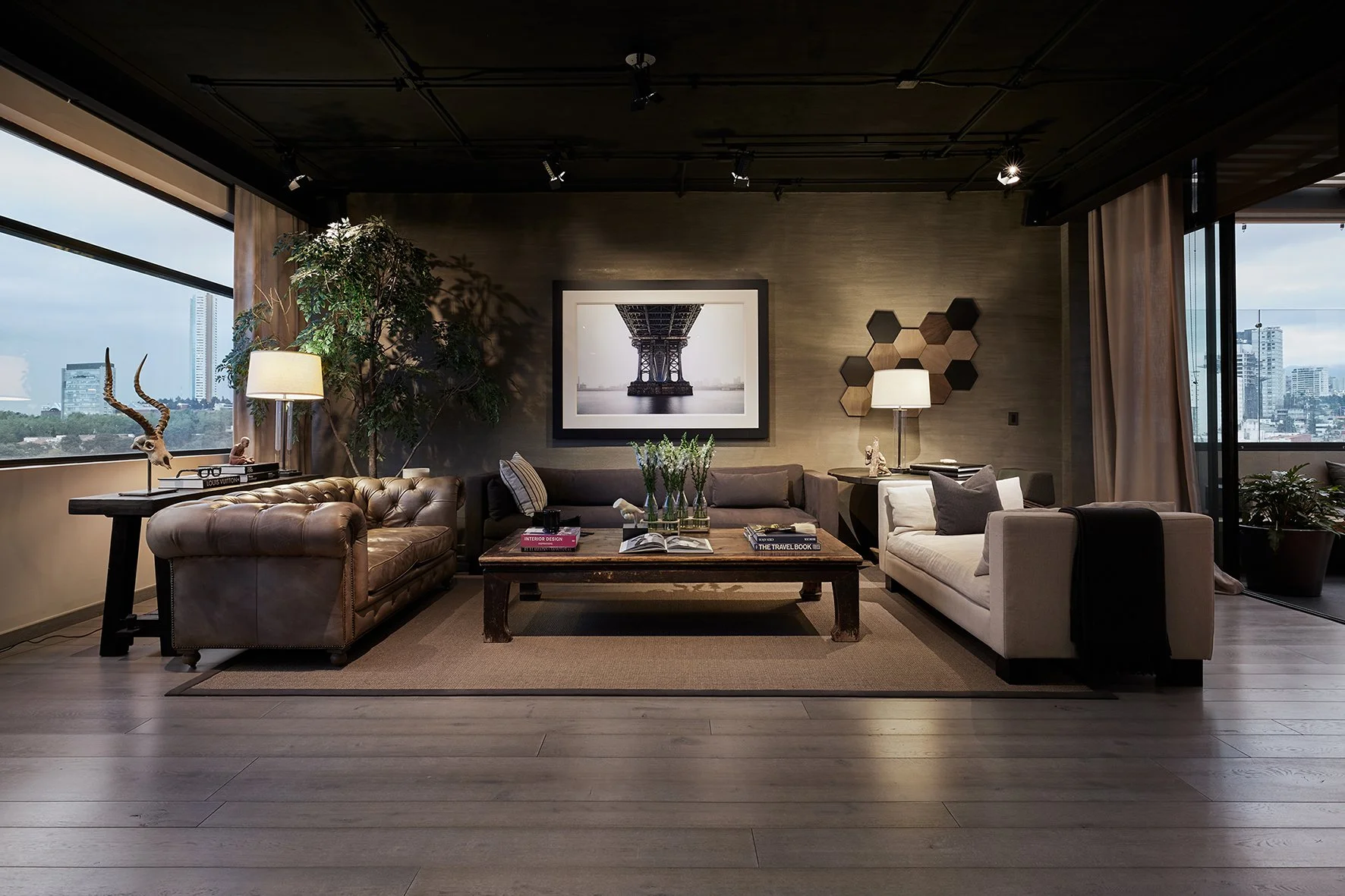 Sala de estar moderna con sillones beige y gris, mesa de café de madera con libros y flores, decoración con cuadros y plantas, grandes ventanas con vista a la ciudad