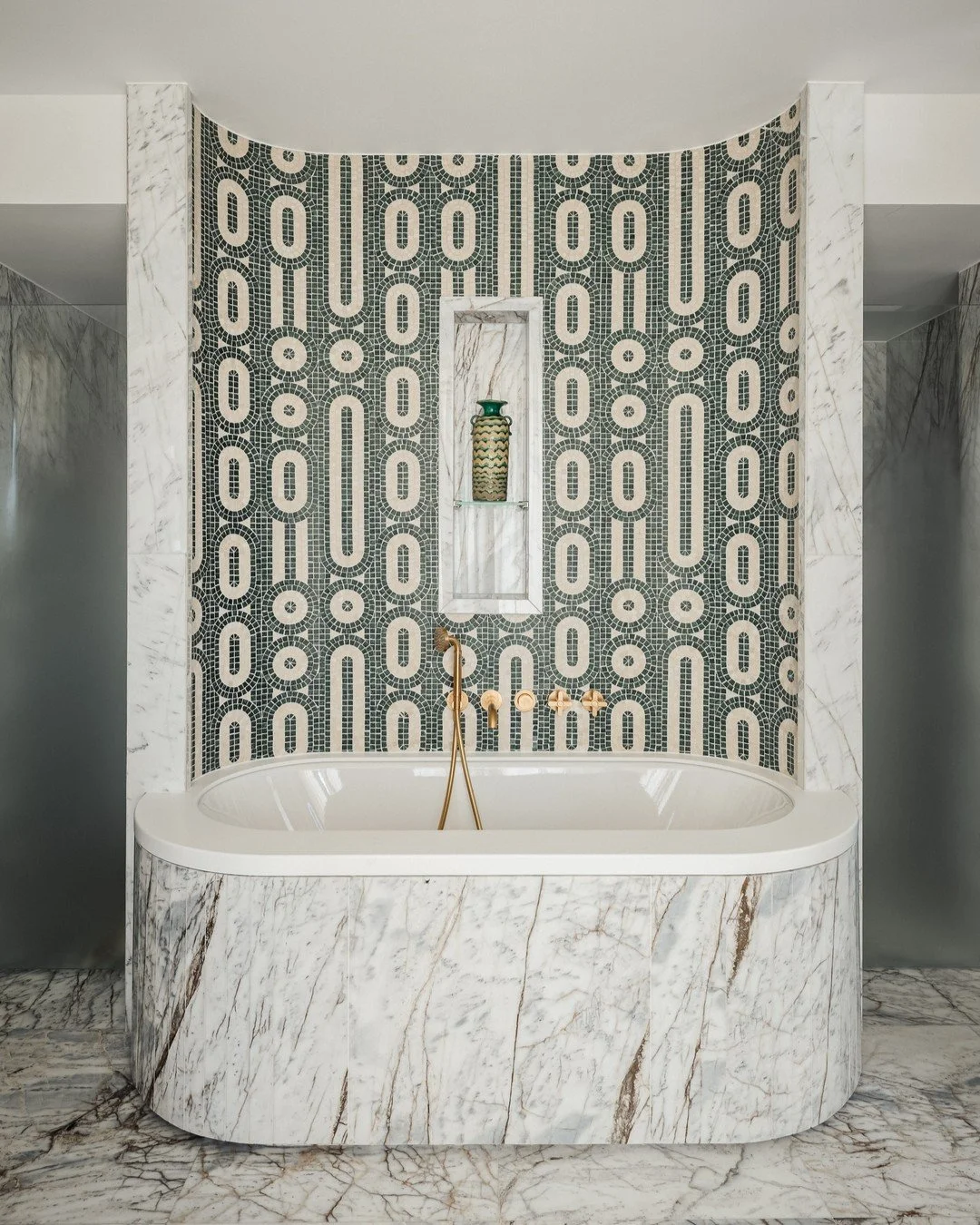 Baño con bañera de mármol blanco, pared de mosaico en tonos oscuros y claros, con un nicho decorativo que contiene una pieza de cerámica y una taza, y piso de mármol con vetas marrones.