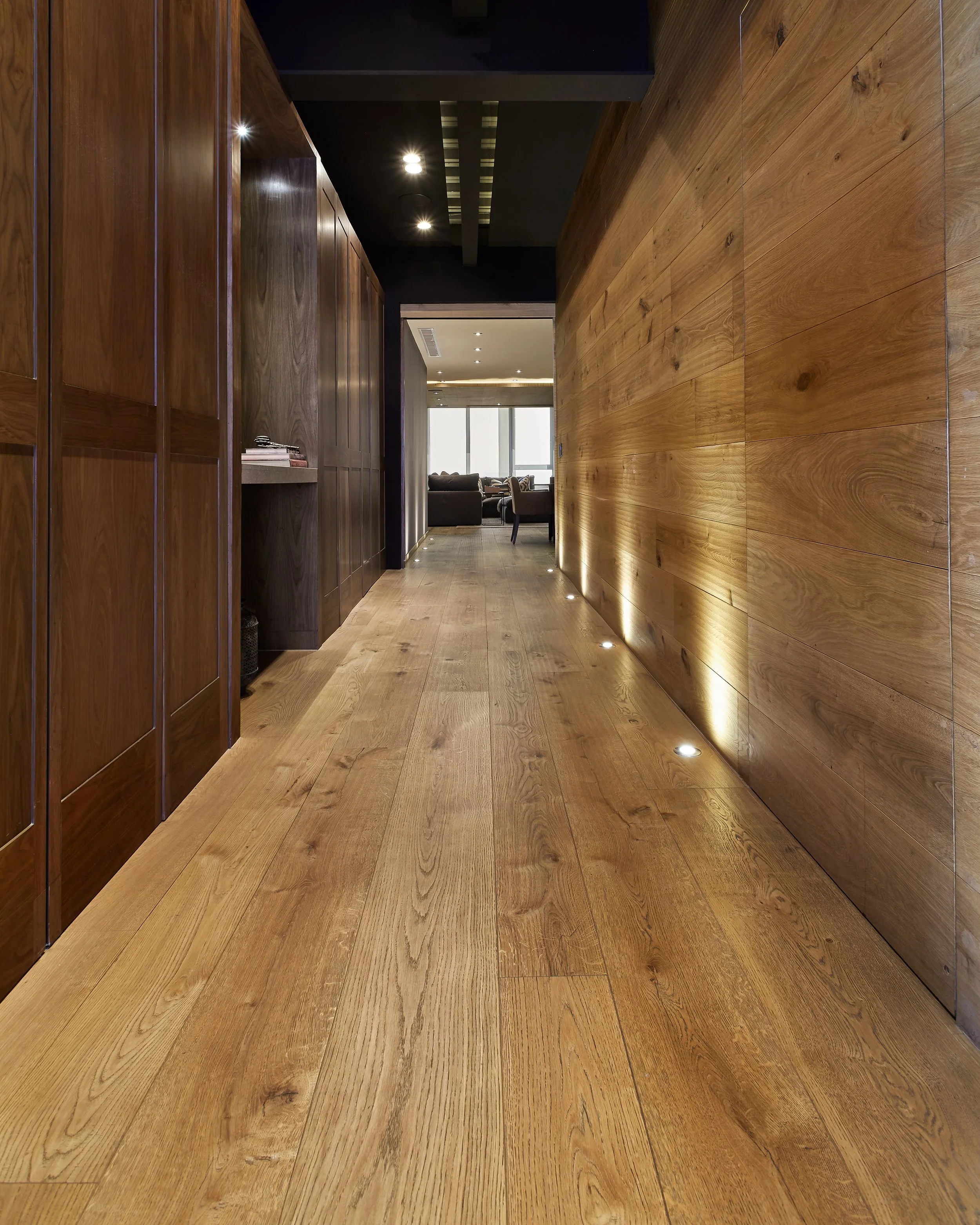 Pasillo con paredes y pisos de madera, iluminación empotrada en el suelo, y una sala de estar con ventanas al fondo.