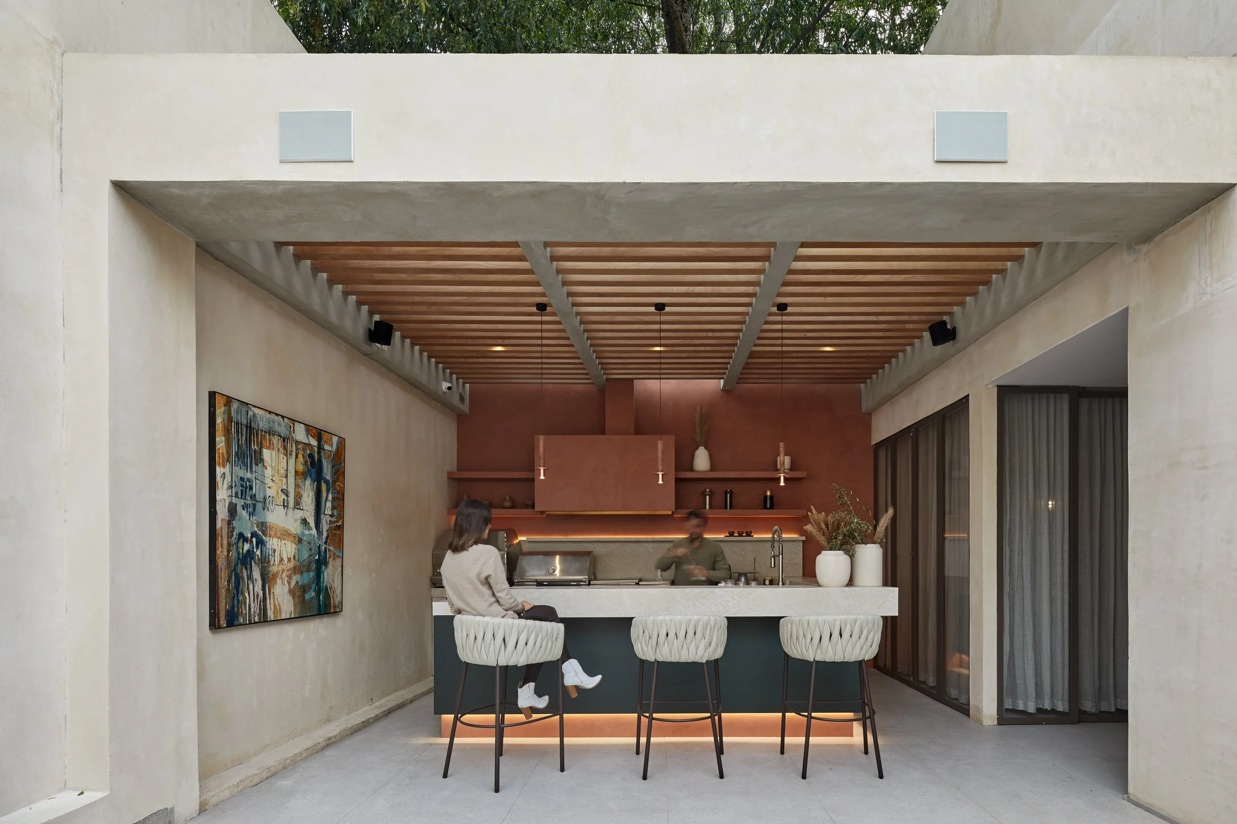 Bar con dos clientes sentados en altares, una mujer y un camarero atendiendo, decoración moderna con paredes en tonos terracota, arte abstracto en la pared, lámparas colgantes y jarrones con plantas decorativas.
