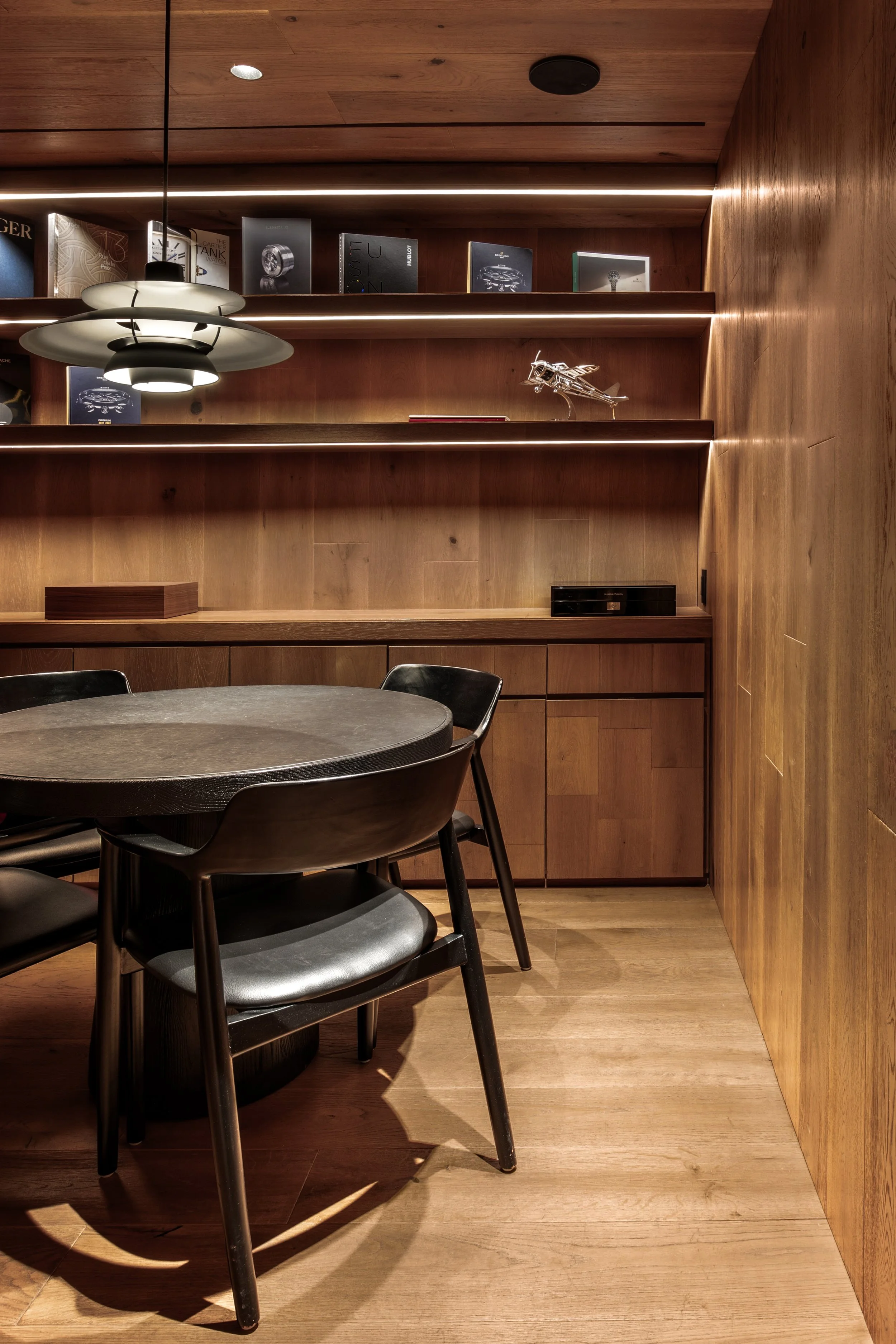 Una sala de estar con paredes, techo y mobiliario de madera, una mesa redonda negra con cuatro sillas negras, y estanterías con libros y una maqueta de avión en la pared.