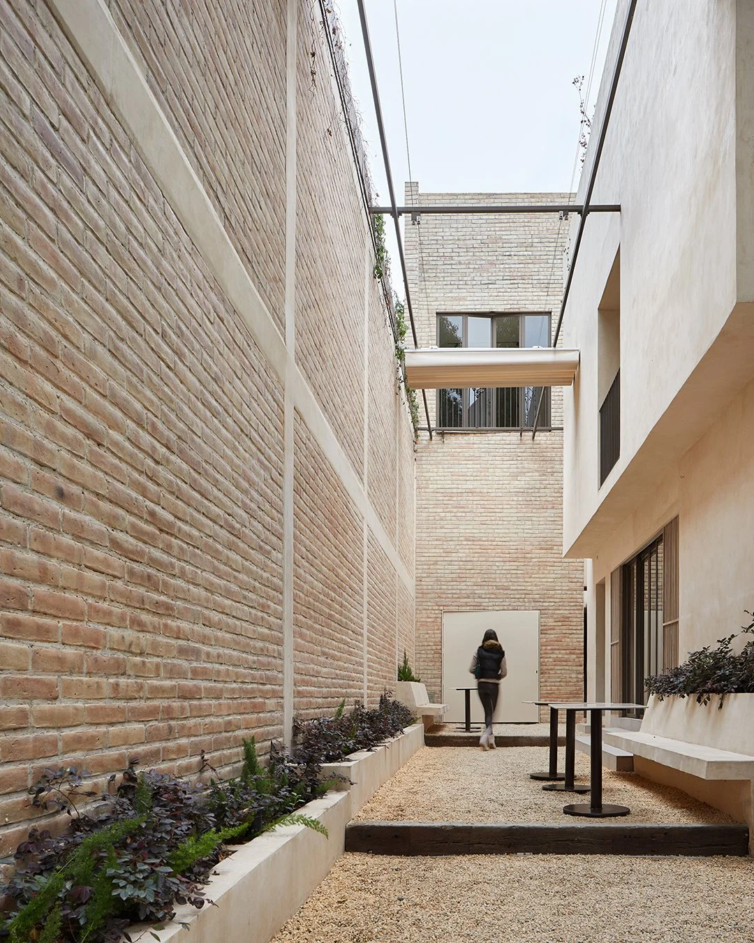 Persona caminando en un patio interno de edificio con paredes de ladrillo, plantas y mesas pequeñas.