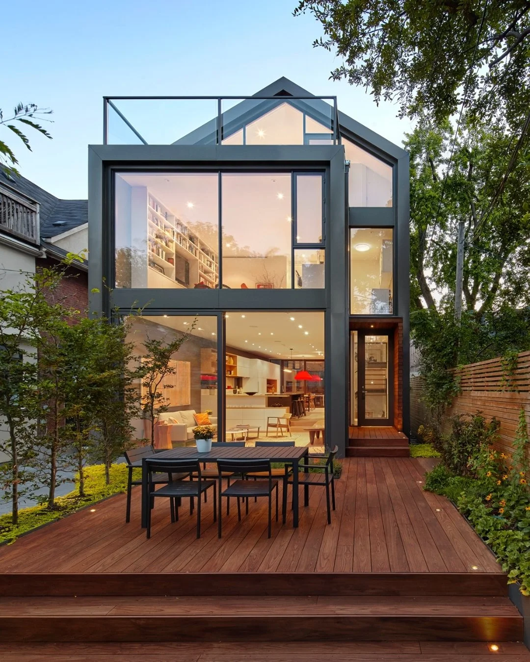 Casa moderna de varios pisos con ventanas grandes, interior visible, jardín de madera con mesa y sillas, árboles y cielo despejado