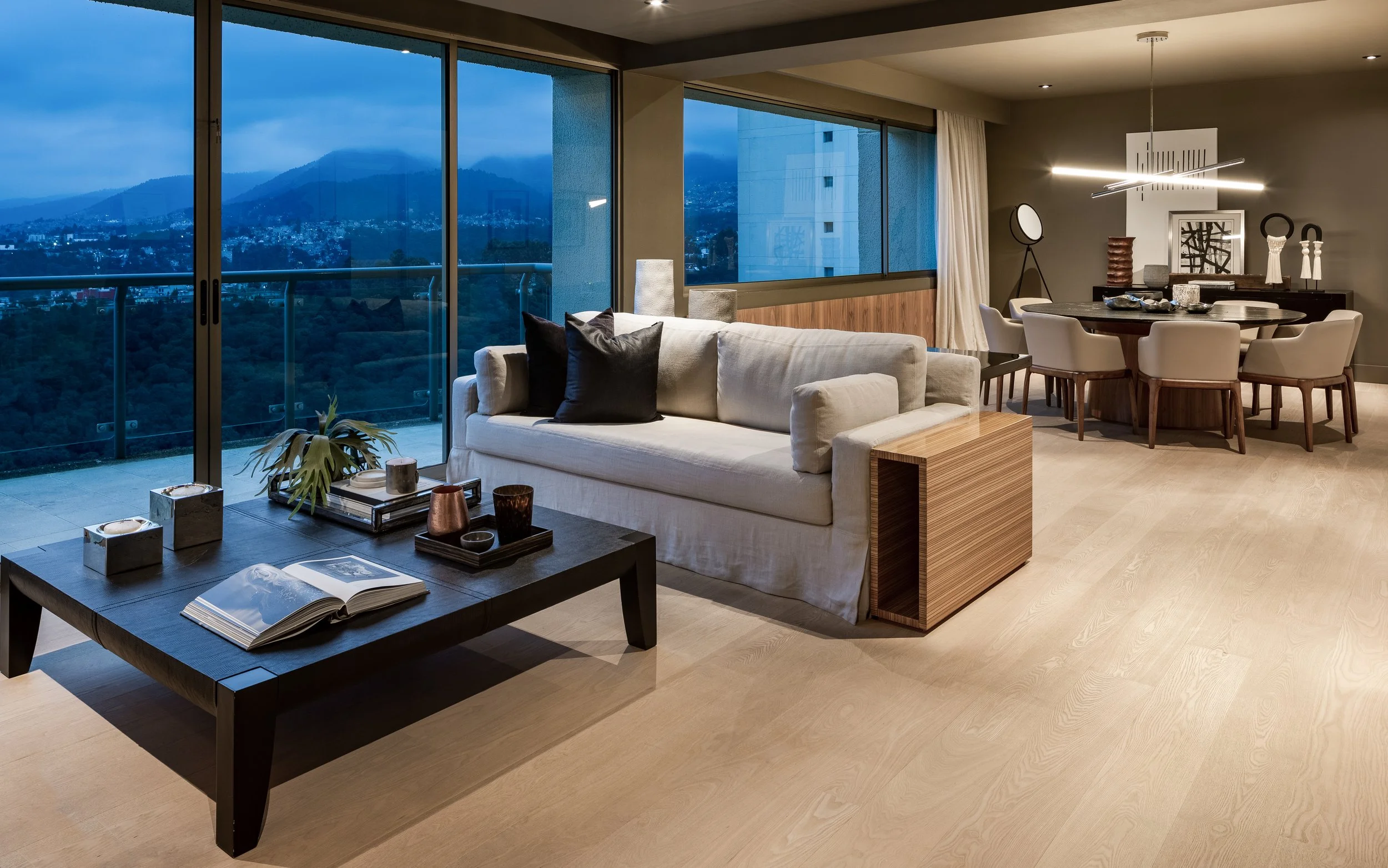 Sala moderna con vistas a la ciudad y las montañas, con sofá blanco, mesa de café negra con libros y objetos decorativos, zona de comedor con varias sillas y decoraciones artísticas.