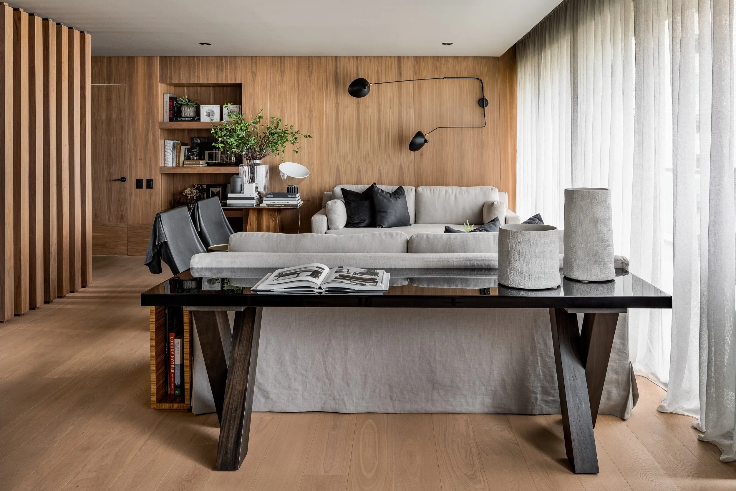 Sala moderna con paredes de madera, sofá blanco con cojines negros, mesa con libros y decoraciones, cortinas blancas en ventana grande.