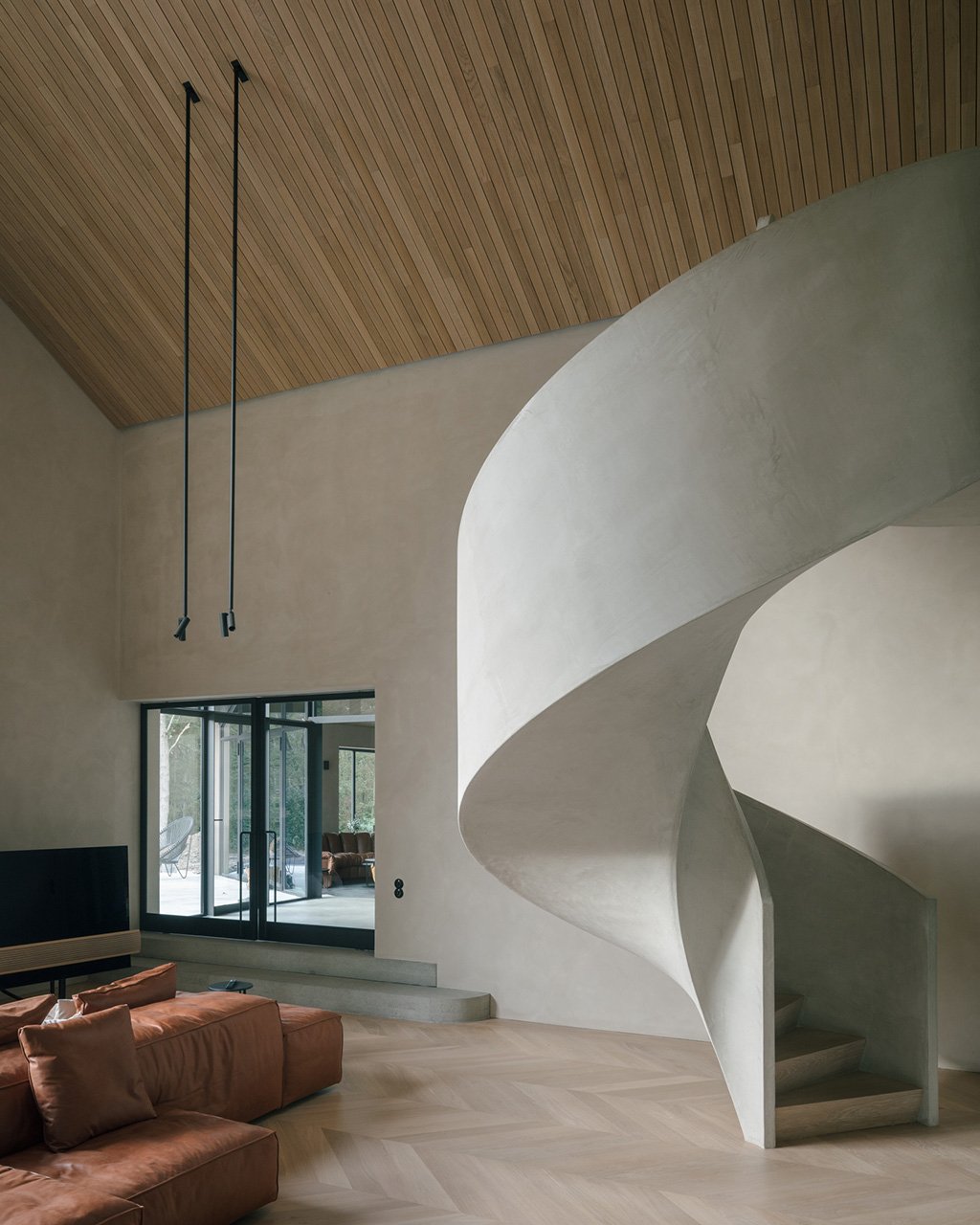 Interior moderno con escalera de espiral de concreto, pared de color claro, techo de madera y puertas de vidrio, en una sala decorada con sofá de color terracota.