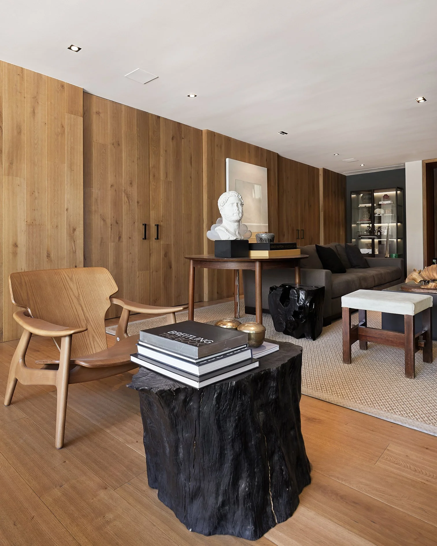 Sala de estar moderna con muebles de madera, sofá gris, mesa rústica con libros y objetos decorativos, paredes de madera y piso de madera clara.