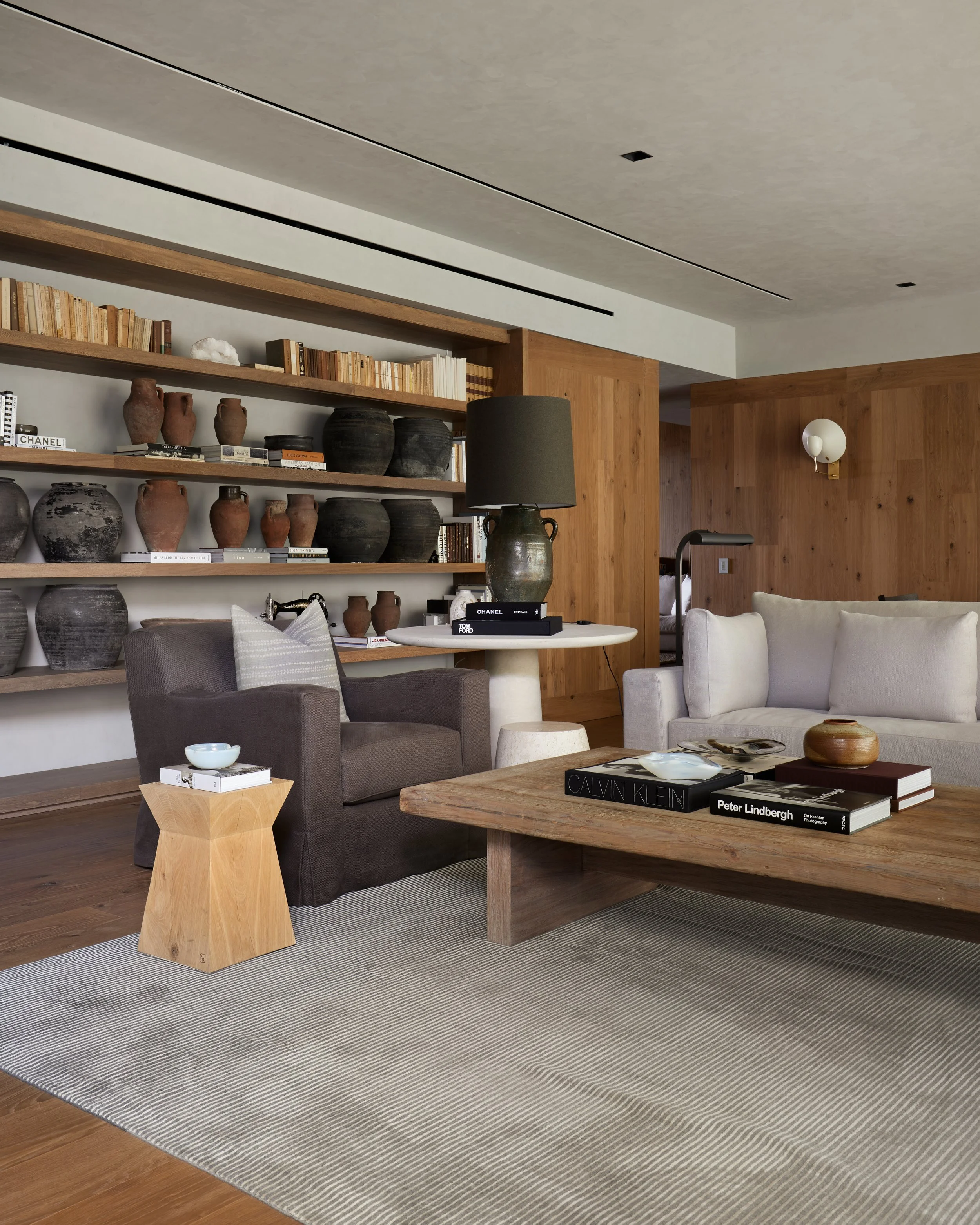 Sala moderna con librero de madera, jarrones y libros decorativos, sofá gris y blanco, mesa de madera con libros y objetos decorativos, alfombra y paredes de madera en fondo.