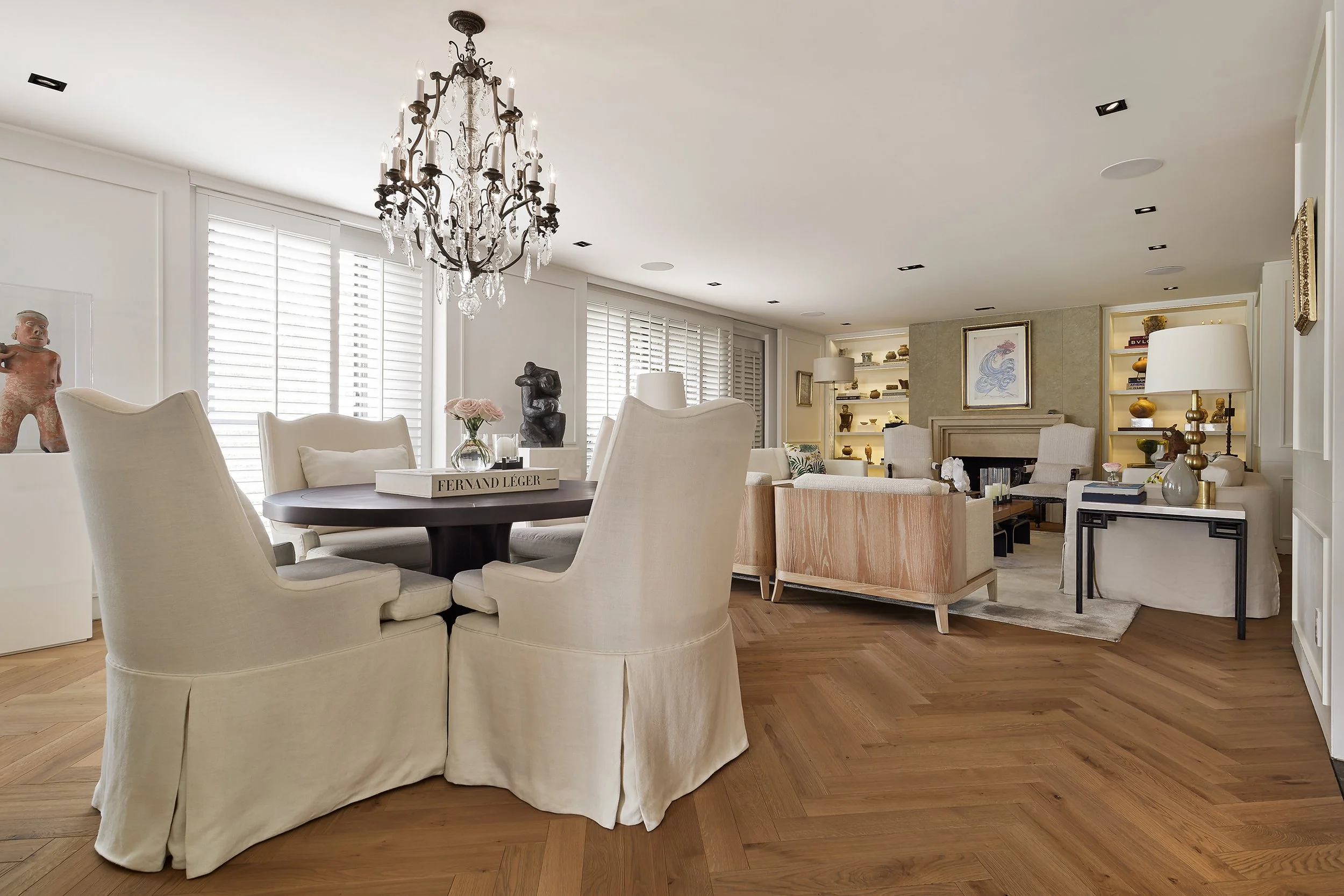 Sala de estar elegante con muebles en tonos claros, chimenea y estanterías con objetos decorativos, iluminada con lámparas de mesa y luz natural de ventanas con persianas. Incluye una mesa redonda con sillas tapizadas y una lámpara de araña
