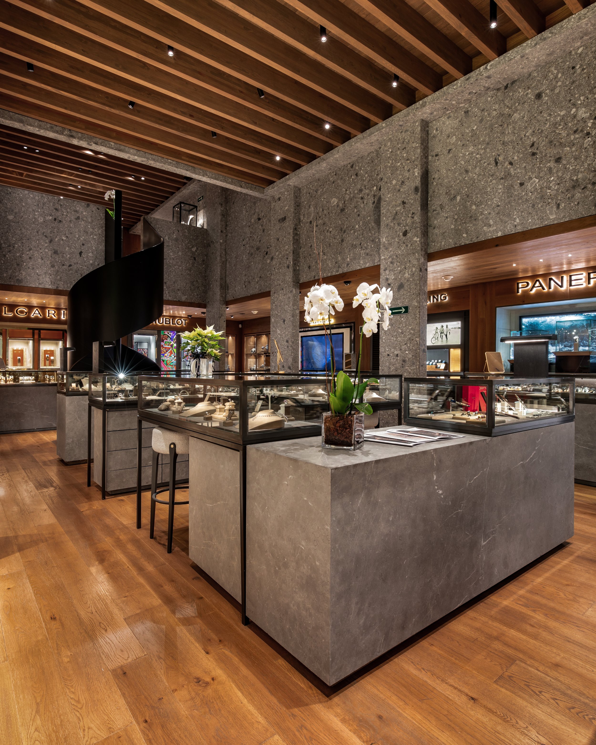 Interior de una joyería con vitrinas de exhibición de relojes y joyas, paredes de piedra gris, suelo de madera, techo de madera con luces empotradas, en el fondo se ven tiendas de marcas como Bulgari y Panerai, y una planta de orquídea blanca en una 