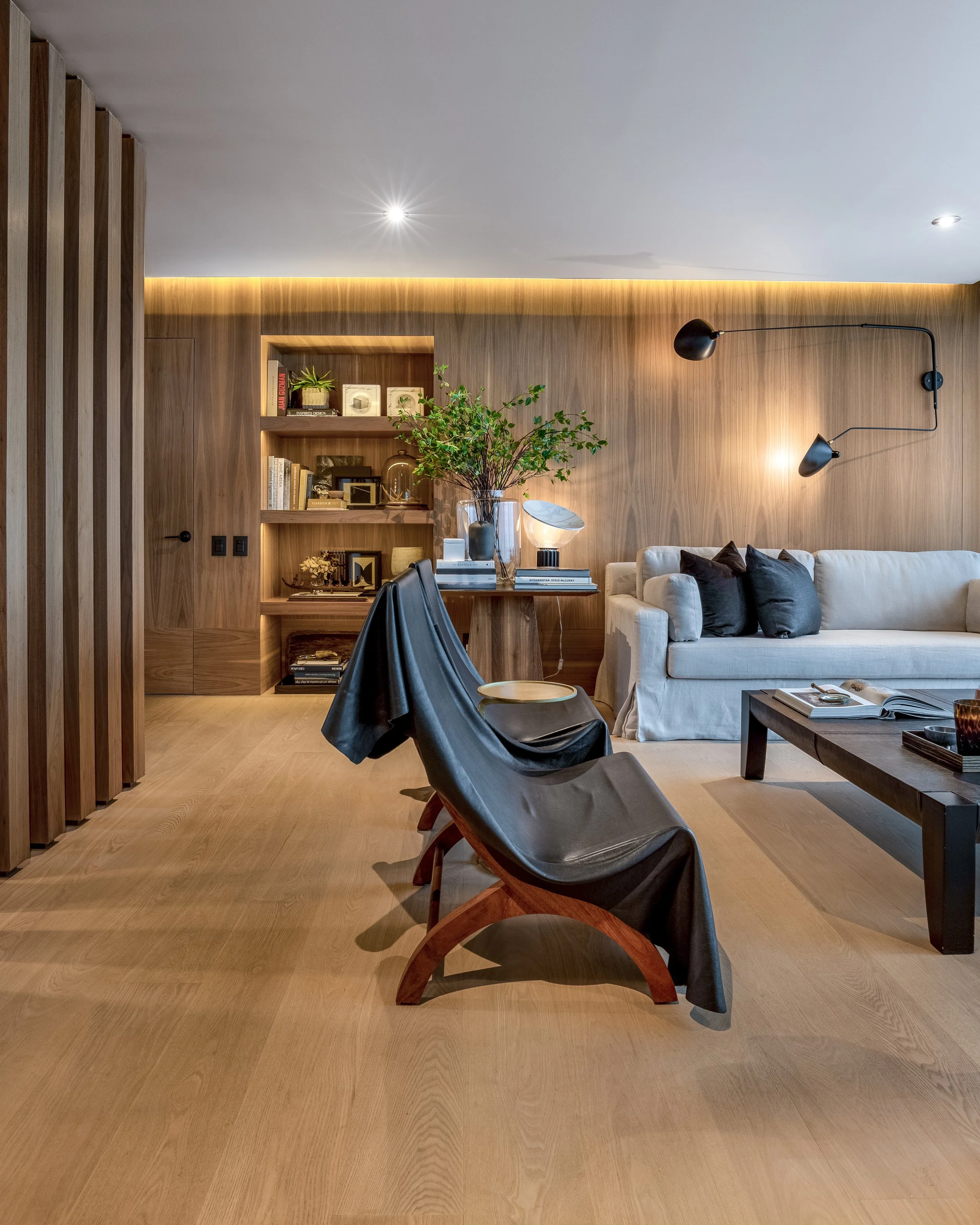 Sala de estar moderna con pared de madera, sofá beige con cojines negros, silla de diseño con manta negra, mesa de centro de madera, estantería empotrada con libros y decoraciones, planta en jarrón y lámparas de pared