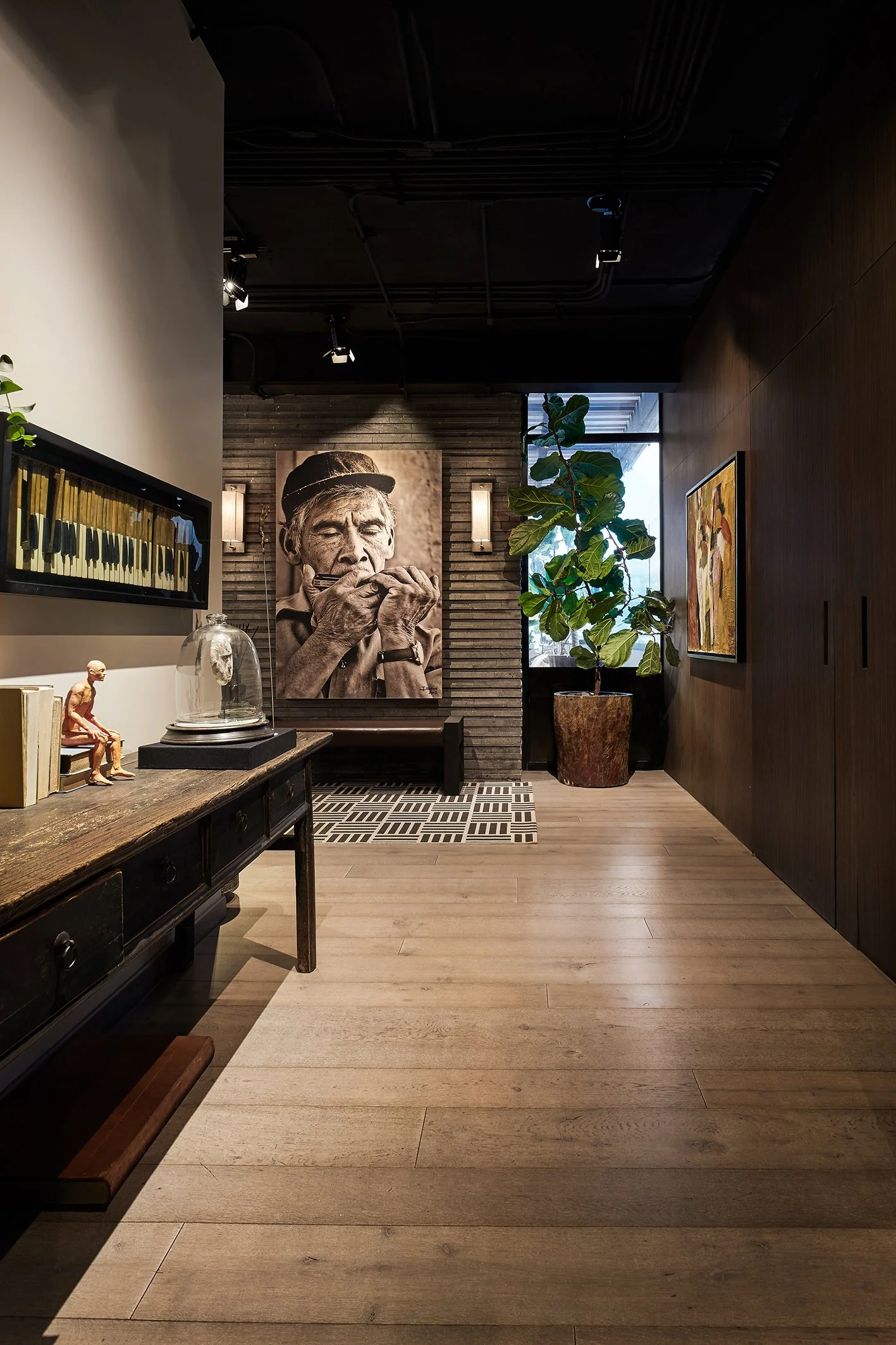 Interior de una sala con madera oscura y decoraciones artísticas, incluyendo una gran foto en blanco y negro de una mujer anciana tocando una flauta, una planta en una maceta grande y varias obras de arte en las paredes.