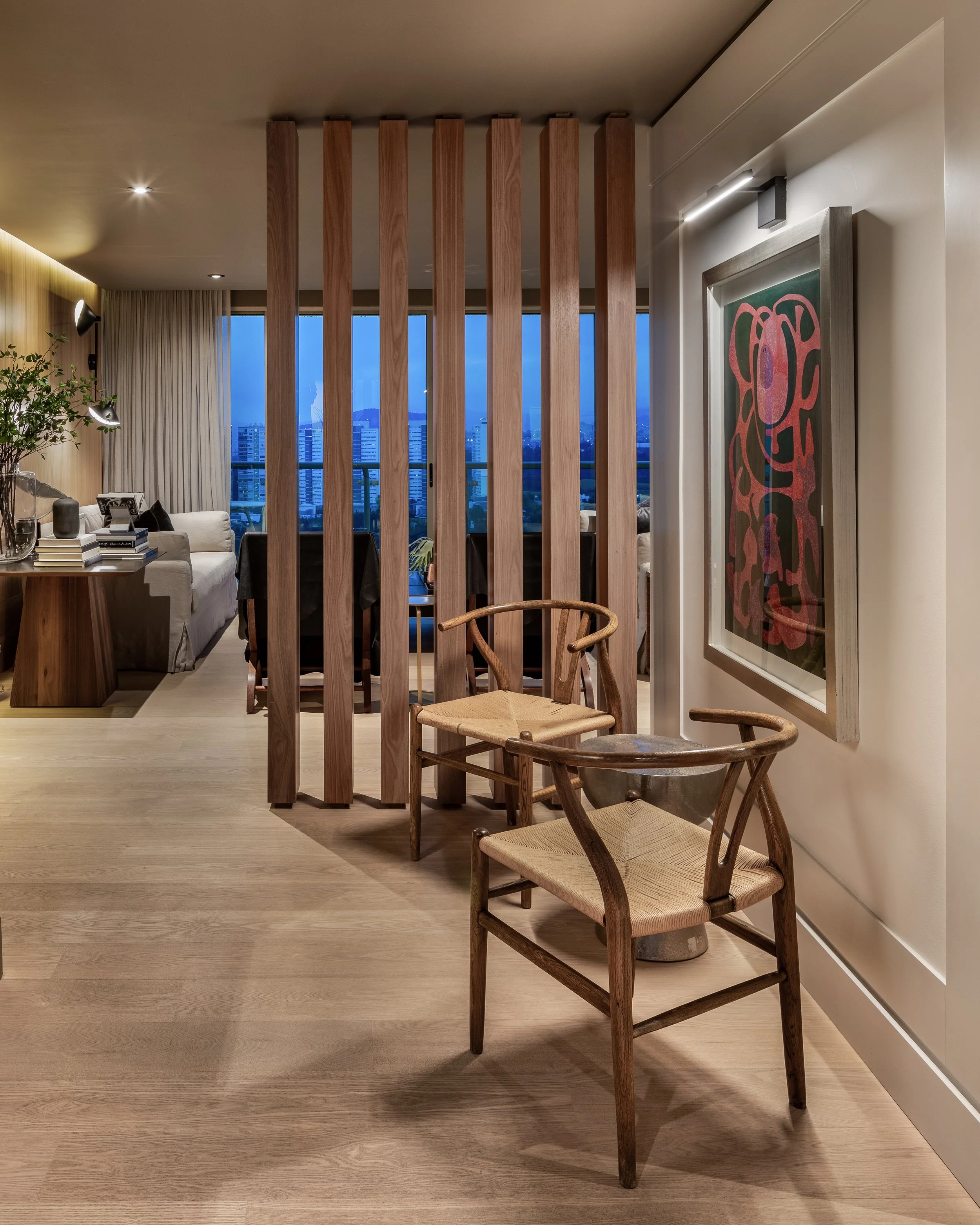 Sala de estar y comedor en un apartamento moderno con vista a la ciudad al atardecer, con muebles de madera y obras de arte en la pared, separadas por un panel de listones de madera.