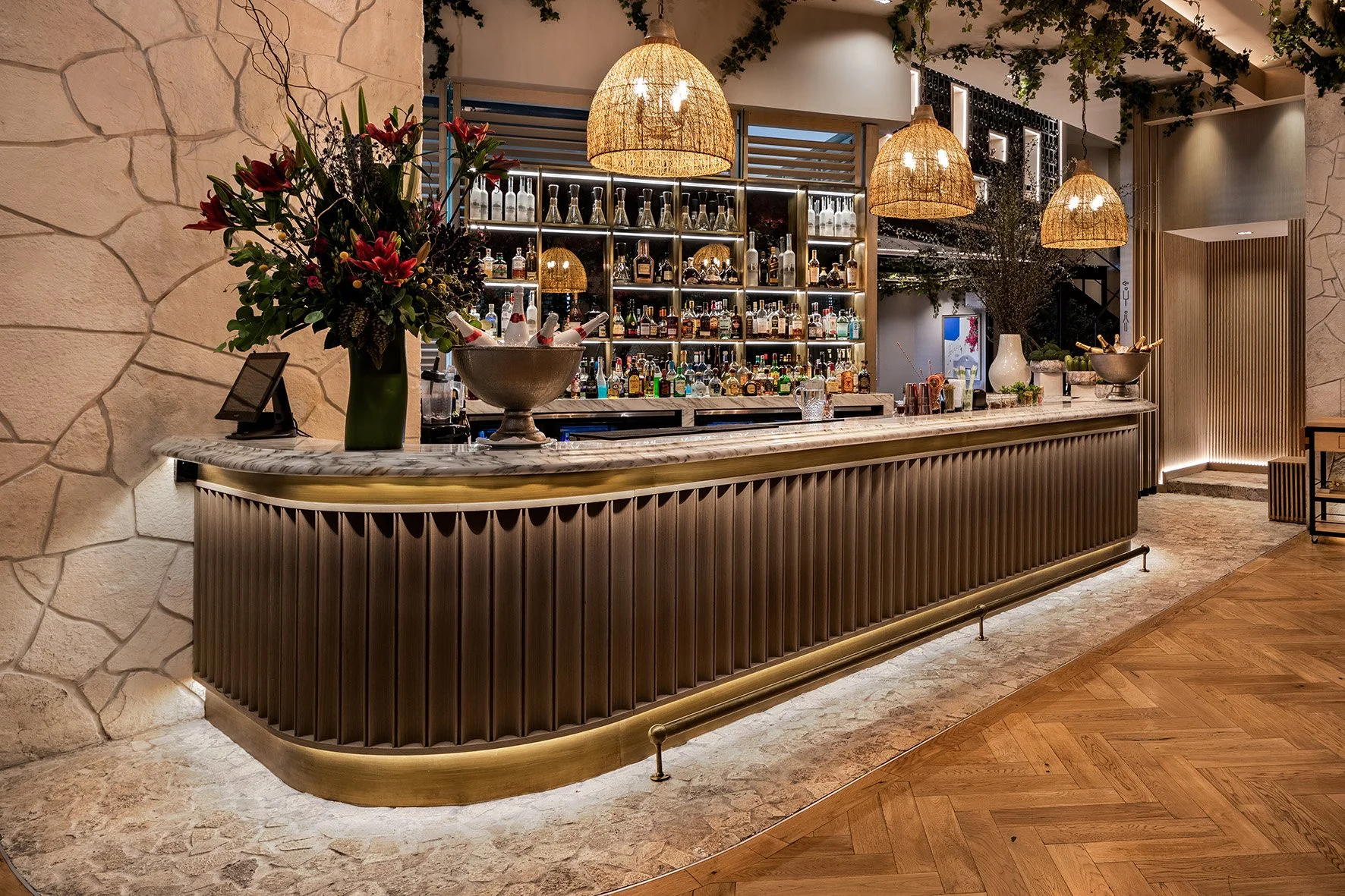Bar con mostrador de mármol y decoración elegante, con estanterías llenas de botellas de licor, luces colgantes de mimbre, flores grandes, y un ambiente acogedor con paredes de piedra y madera.