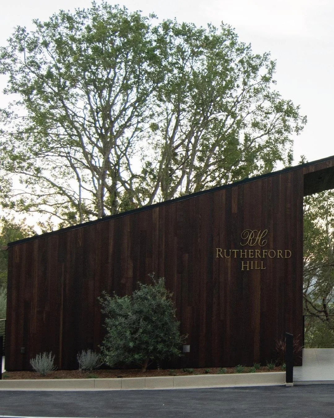 Edificio con fachada de madera oscura y letrero que dice 'DH Rutherford Hill', rodeado de plantas y con árbol grande de hojas verdes detrás.