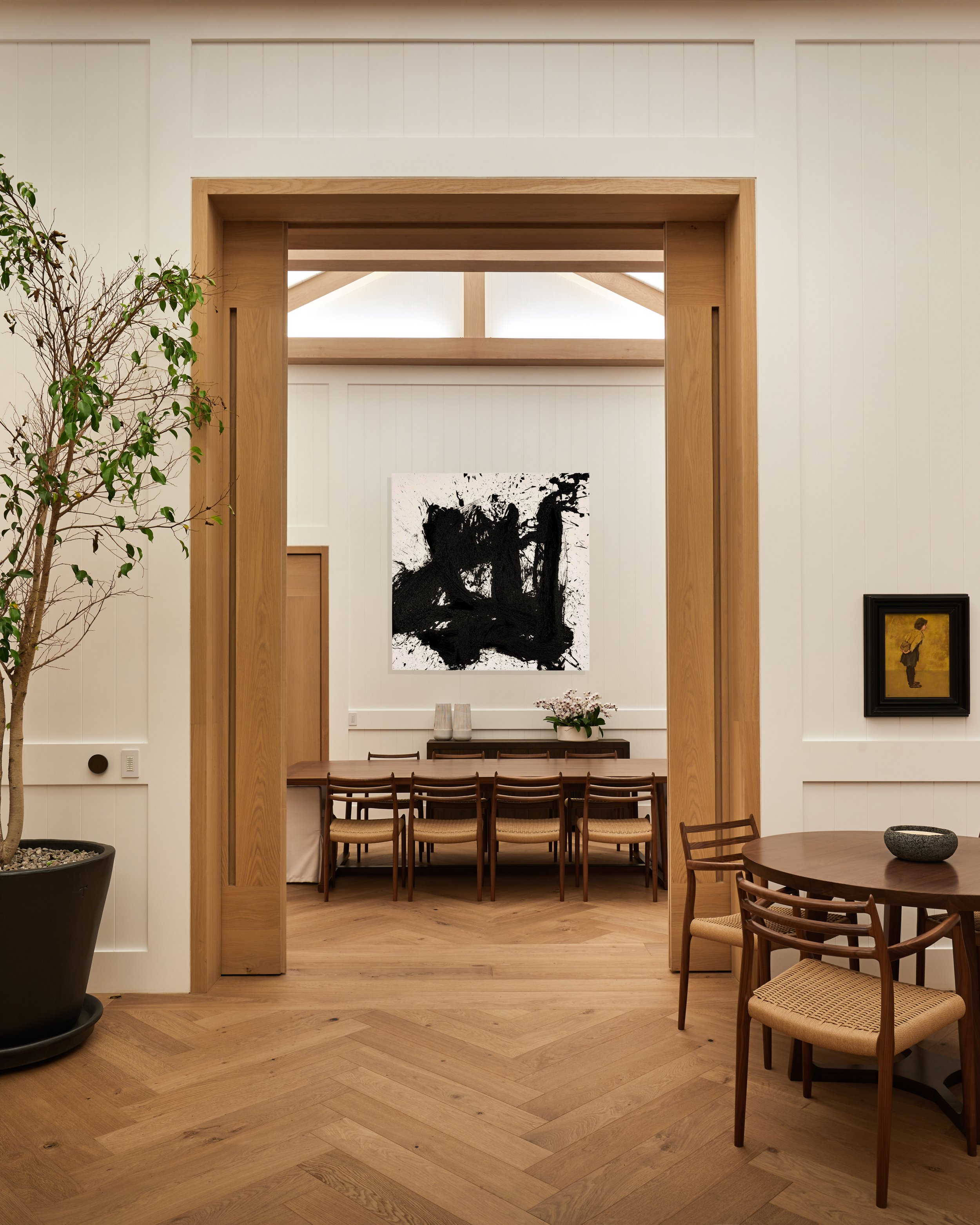 Sala de comedor en una casa con paredes blancas y suelo de madera, decorada con obras de arte modernas y un árbol en una maceta grande.
