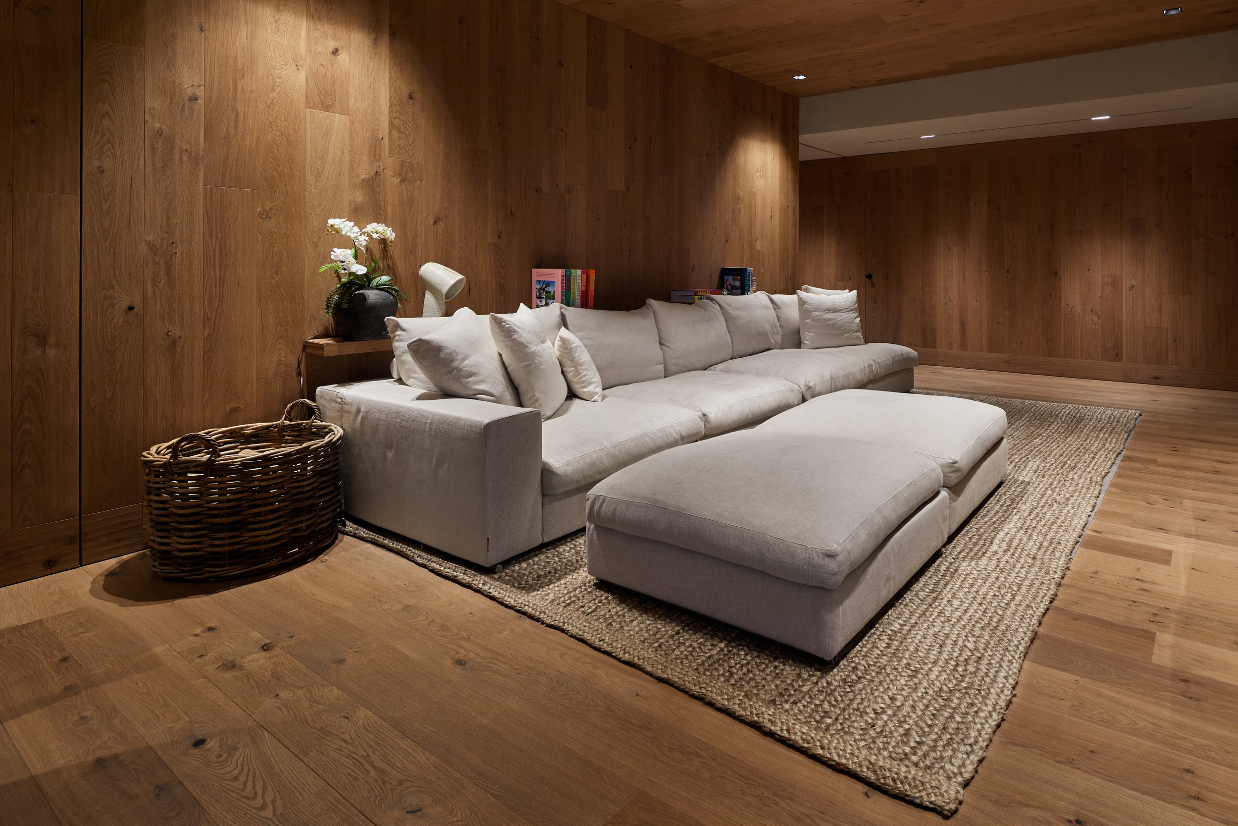 Sala de estar con sofá grande y cojines, en paredes y suelo de madera, con alfombra y decoraciones de plantas y libros.