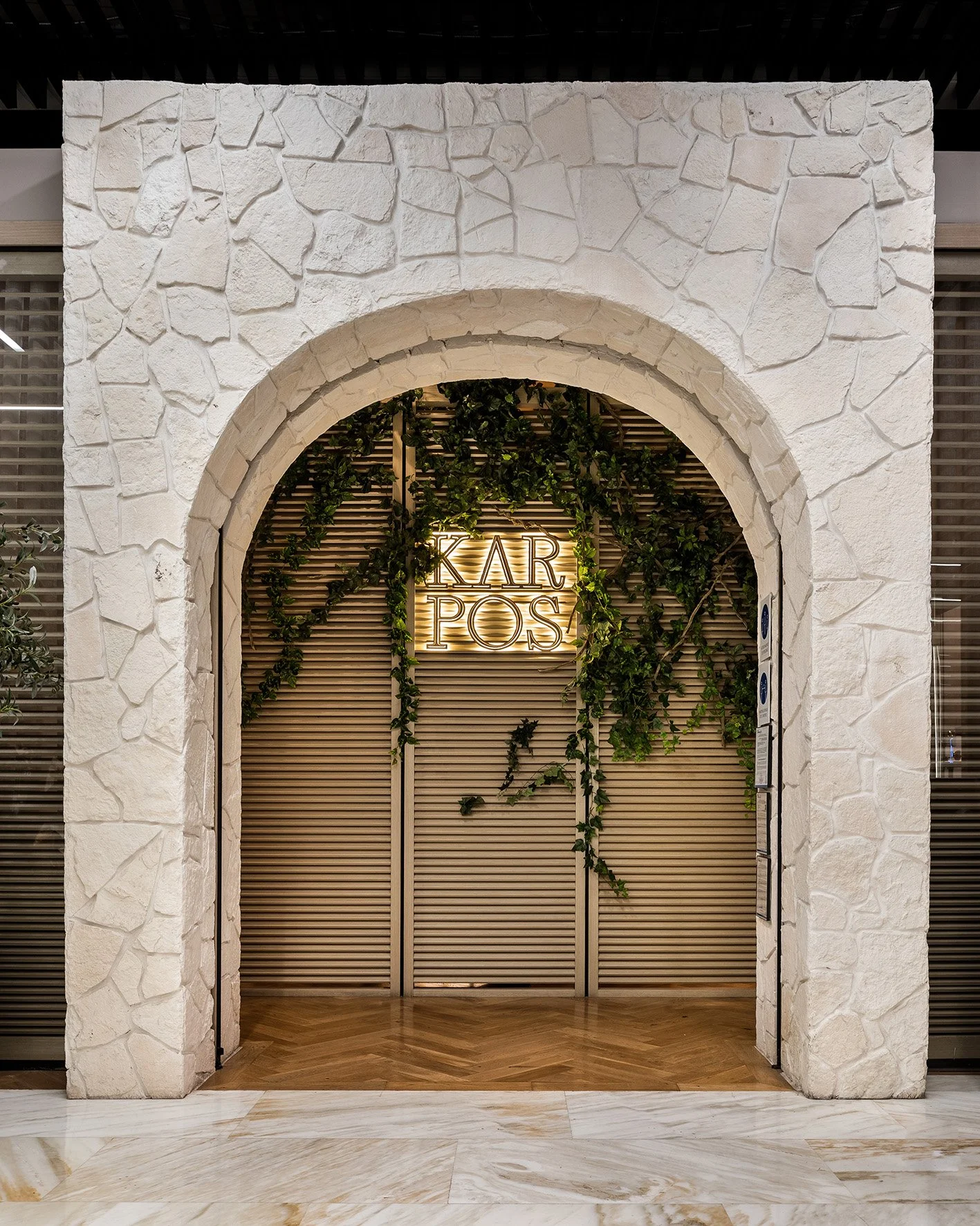 Entrada con arco de piedra blanca, puertas de madera con listones horizontales, follaje y cartel luminoso que dice 'KARPOS'.