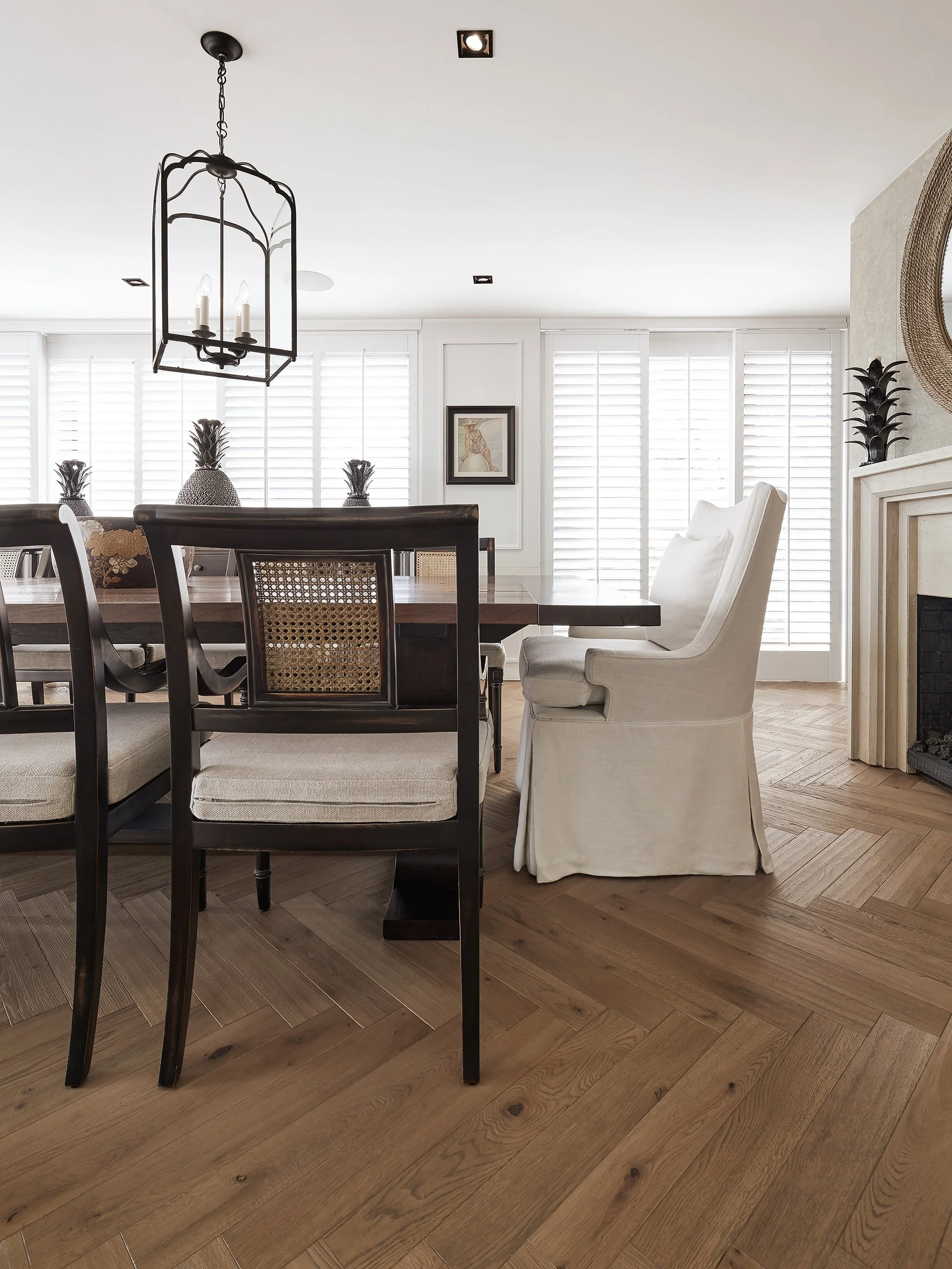 Comedor con sillas de madera oscura, una de ellas blanca con cortinas blancas y ventanas con persianas blancas de madera.