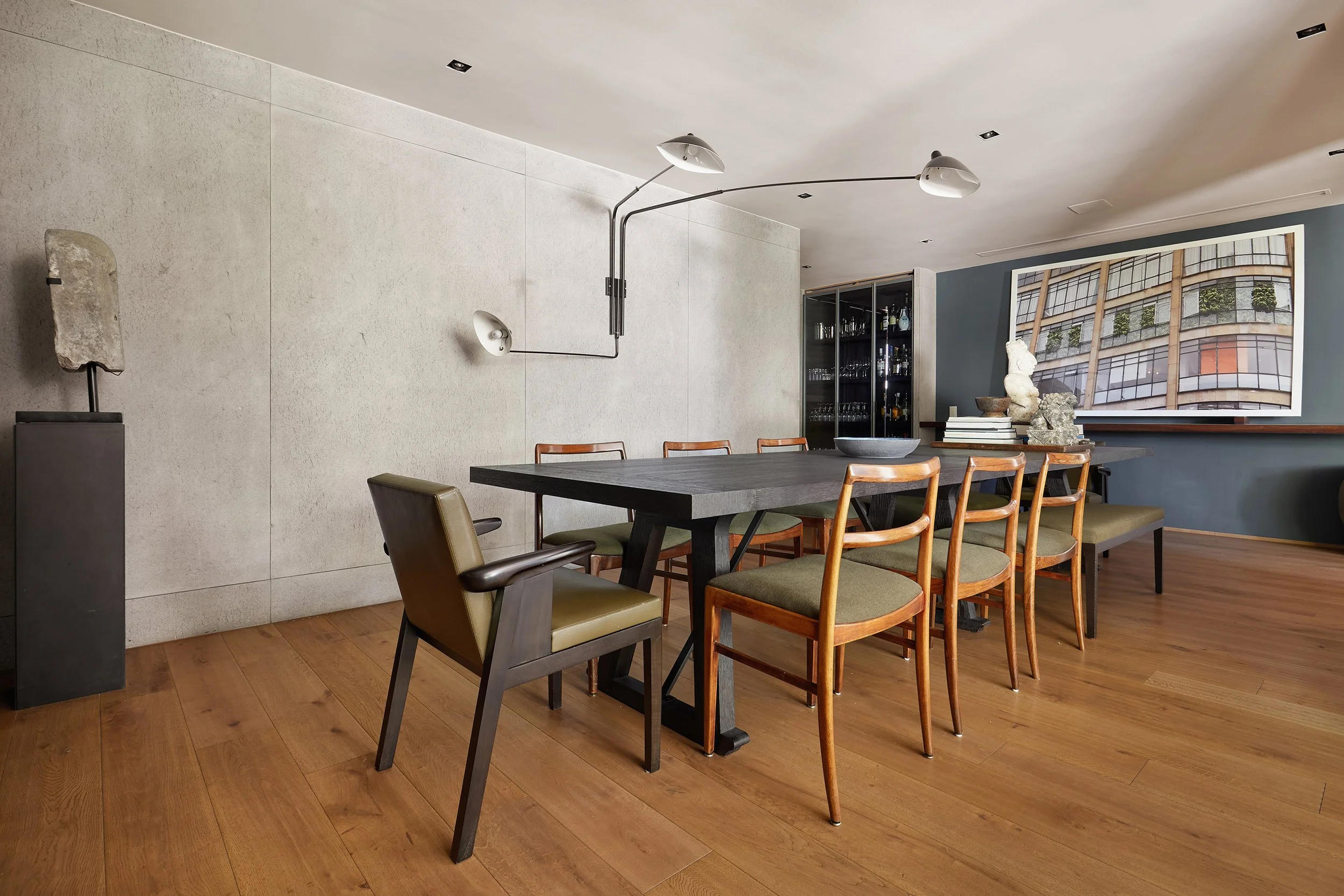Sala de comedor moderna con mesa de madera oscura y sillas de diferentes estilos y colores, incluyendo una silla de oficina y sillas de madera con cojines verdes. En la pared de fondo hay un gran marco con una fotografía de un edificio. Sobre la mesa