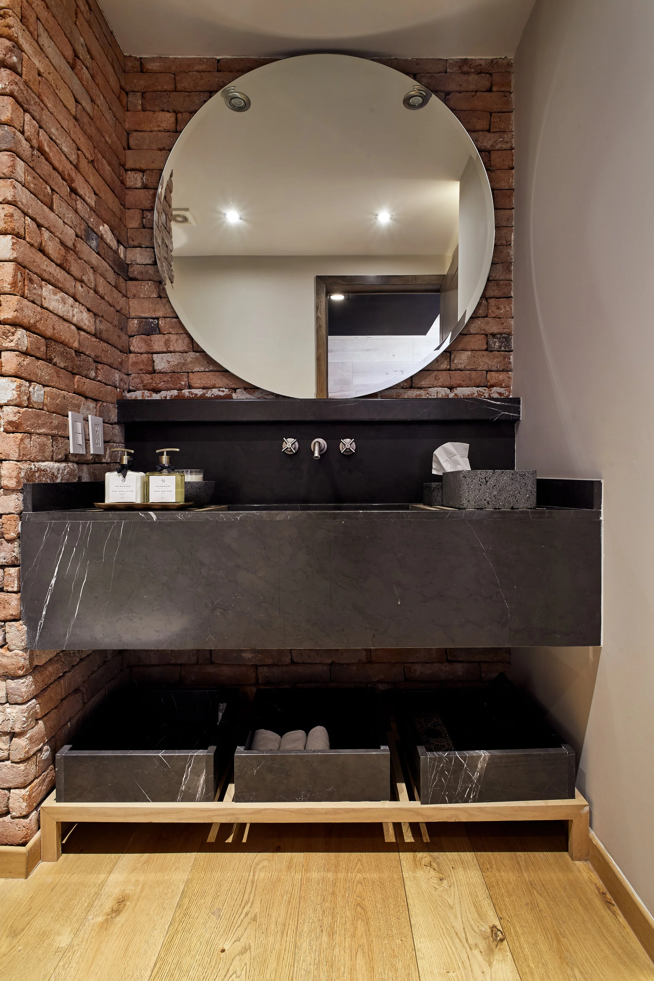 Un lavabo de piedra negra con un espejo redondo y paredes de ladrillo expuesto en un baño moderno.