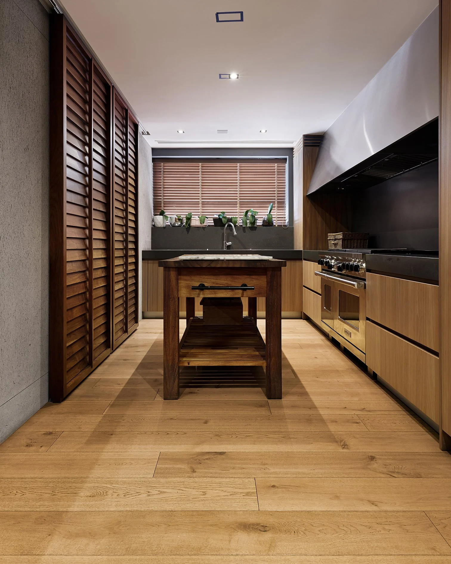 Cocina moderna con madera clara, ventana con persianas de madera, encimera negra, estantes con plantas, estufa con campana de acero inoxidable y mesa de madera en el centro.