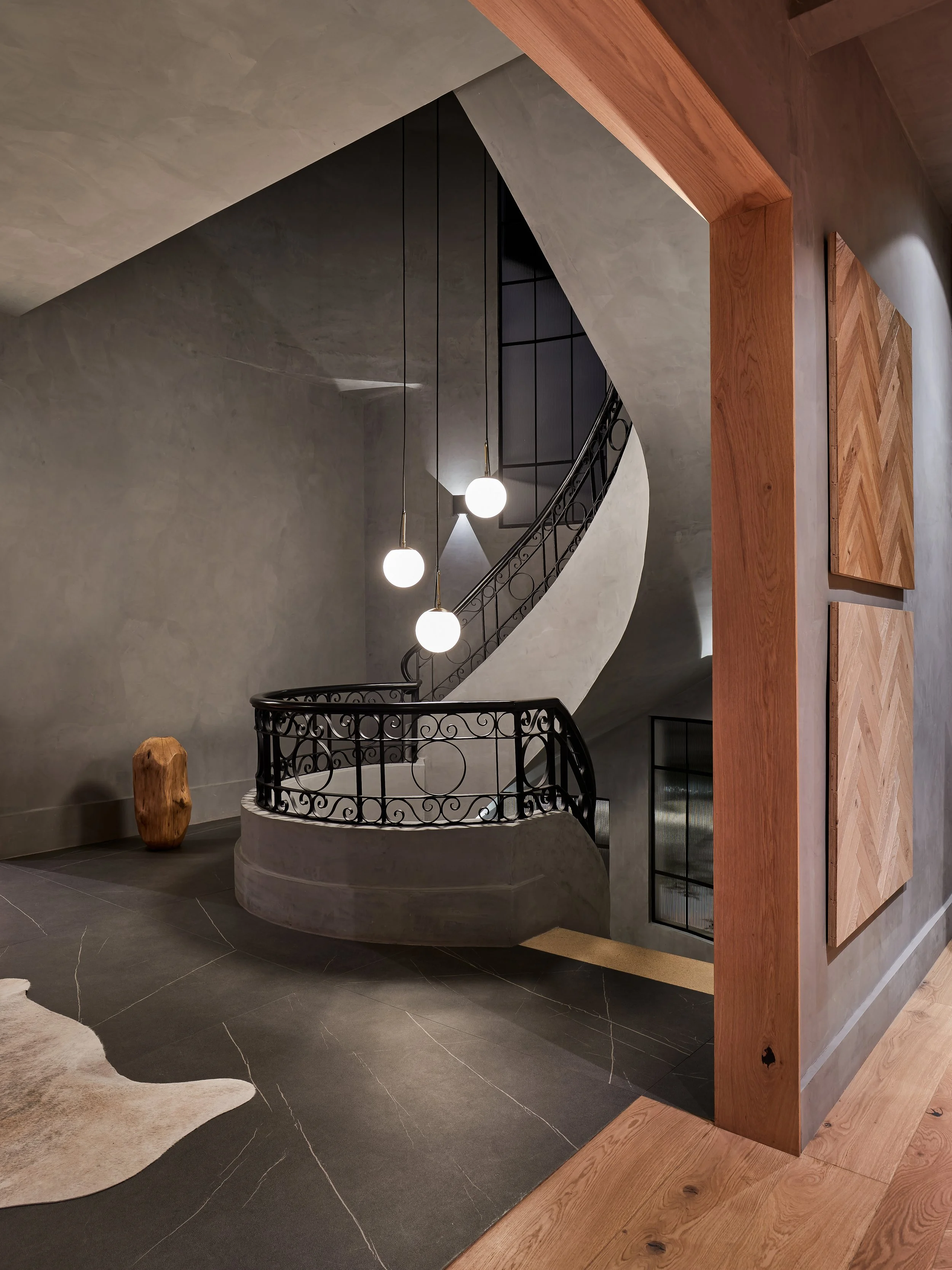 Escalera de vide en un interior moderno con barandilla de hierro decorativa, paredes de cemento y lámparas colgantes de luz blanca, con accesorios de madera y un tapete de piel en el suelo.