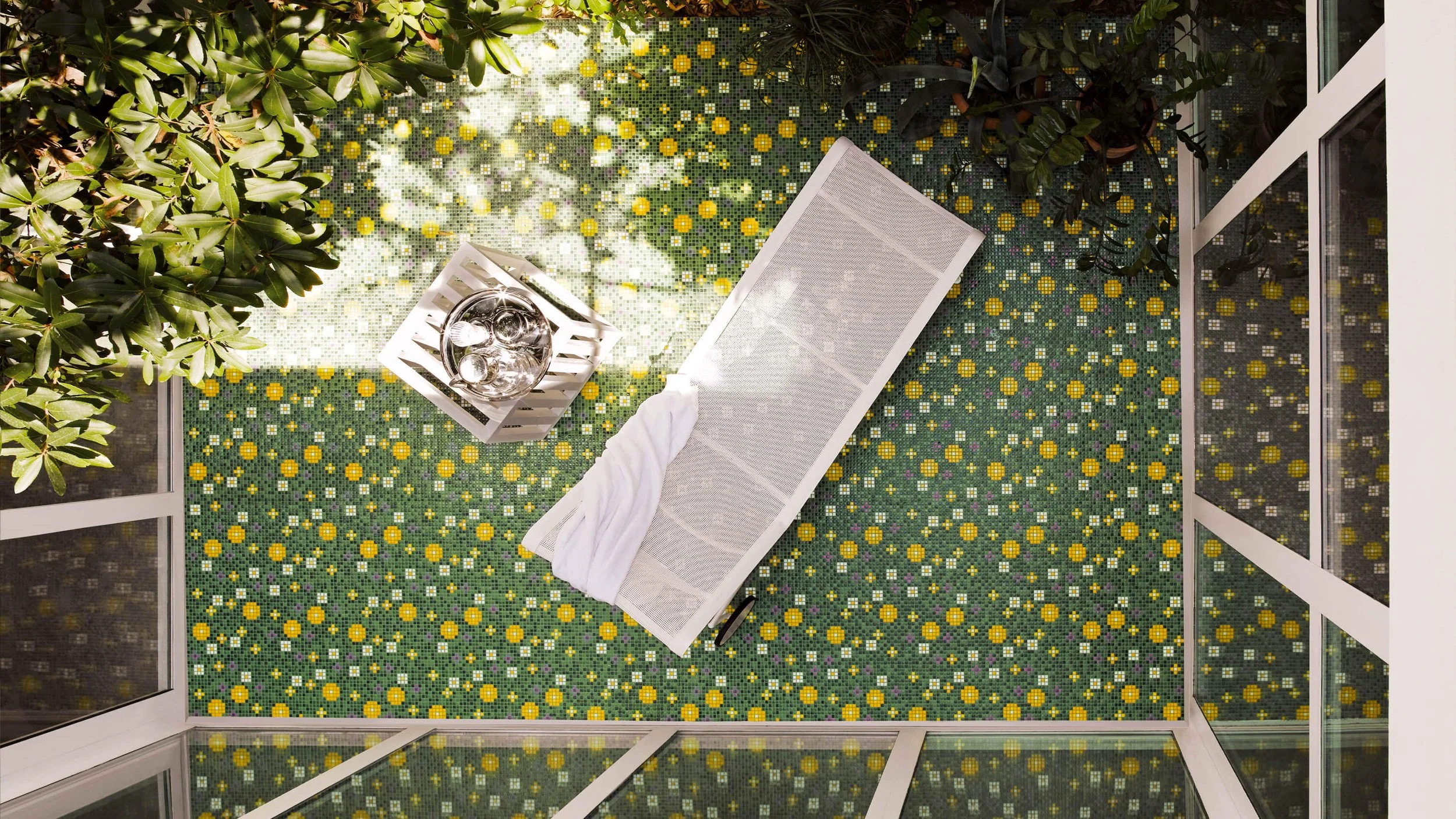 Terraza con piso de mosaico de flores amarillas y verdes, sillón de malla, blanco con cobija y mesa decorativa con vasos de agua y platos de comida, rodeada de plantas verdes y paredes de cristal.