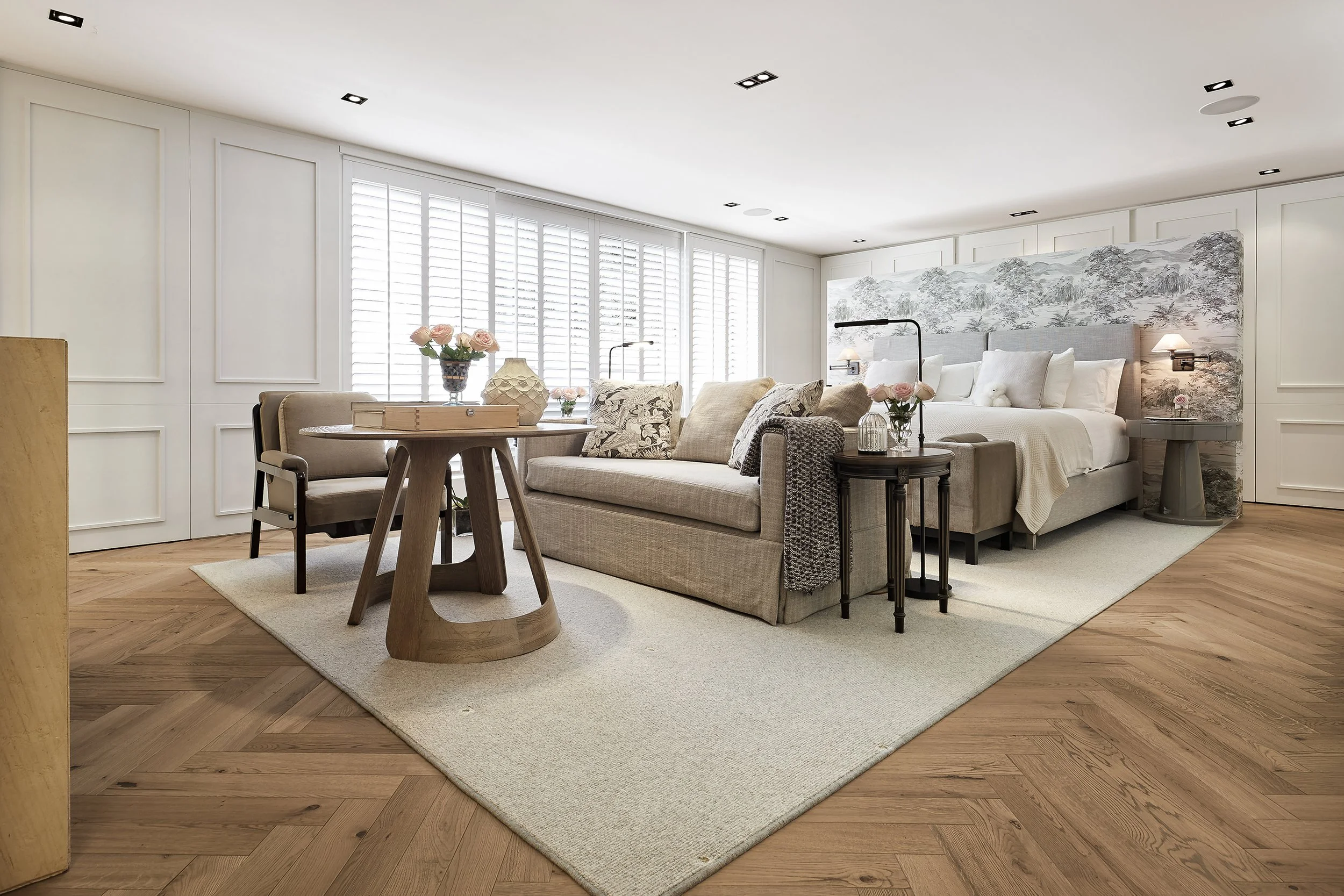 Habitación en tonos neutros con cama grande, sillas, mesa redonda con flores, sofá, alfombra clara y paredes con mural de paisaje en acuarela.