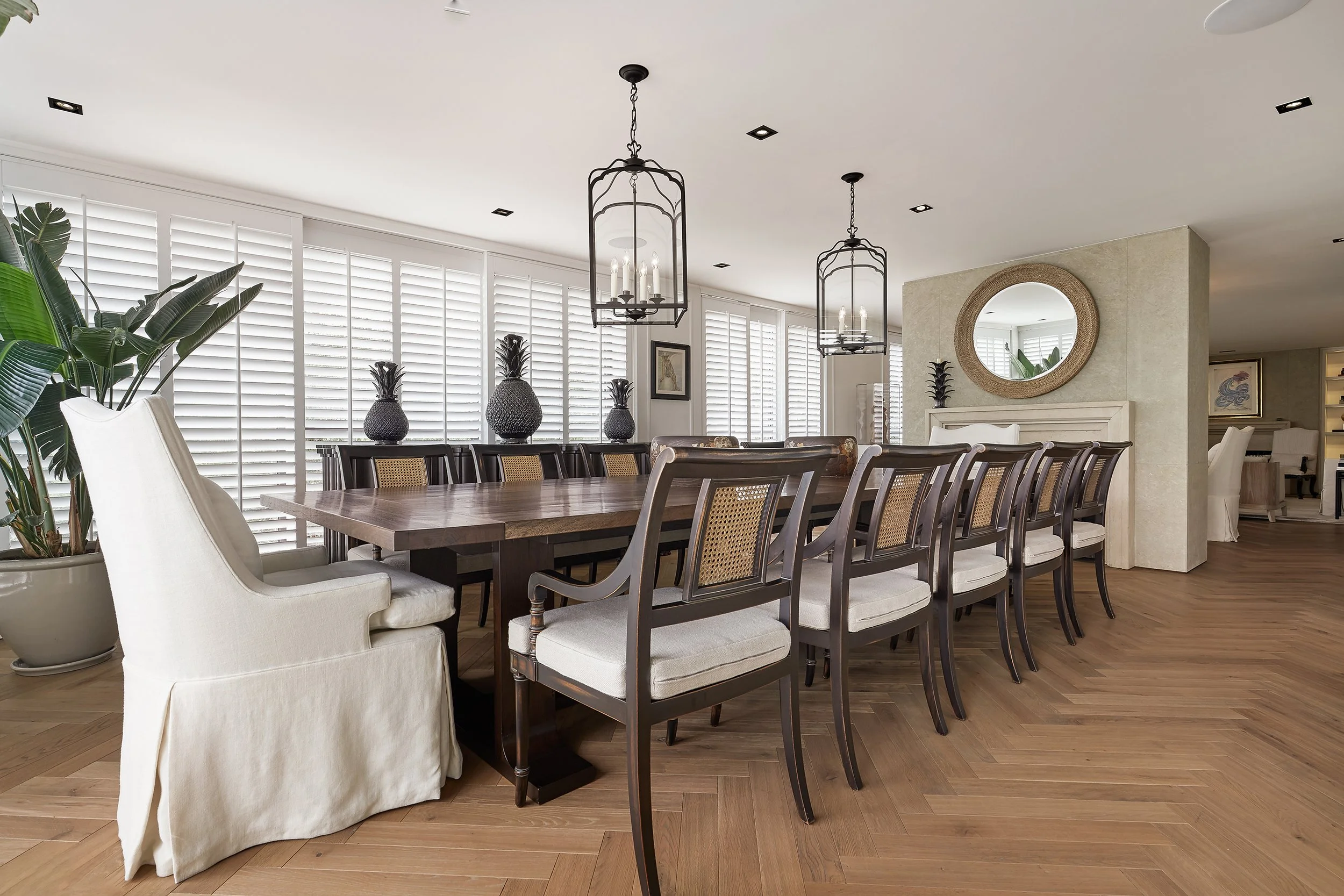 Sala de comedor elegante con una larga mesa de madera, sillas de madera y tapiz beige, lámparas colgantes negras, plantas y decoración moderna con espejo redondo en la pared.